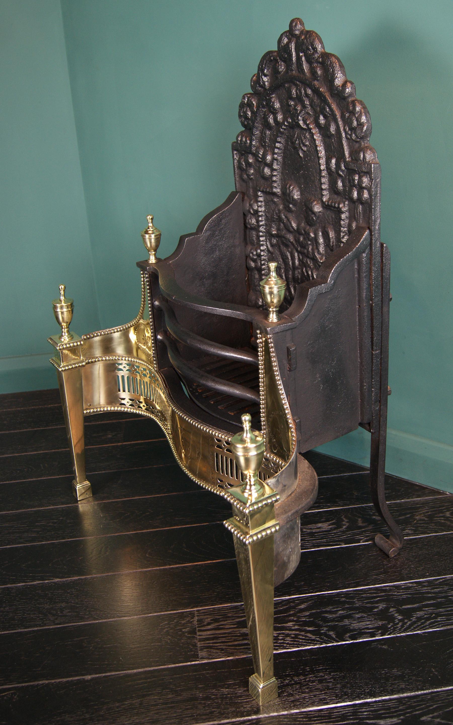 A brass, wrought & cast iron fire grate in the Adam manner by Thomas Elsley. The railed serpentine basket above a polished brass pierced fret centered by an engraved tablet, set between tapering engraved legs, with urn finials. Decorative fireback