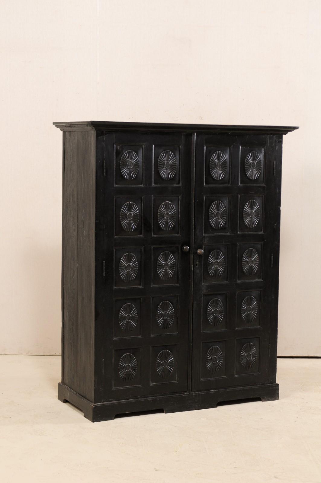 A British Colonial ebonized wood cabinet from the early to mid-20th century. This British Colonial cabinet from India, standing approximately 5.25 feet in height, features two doors, adorn in raised panels with oval/floral centers, and a nicely