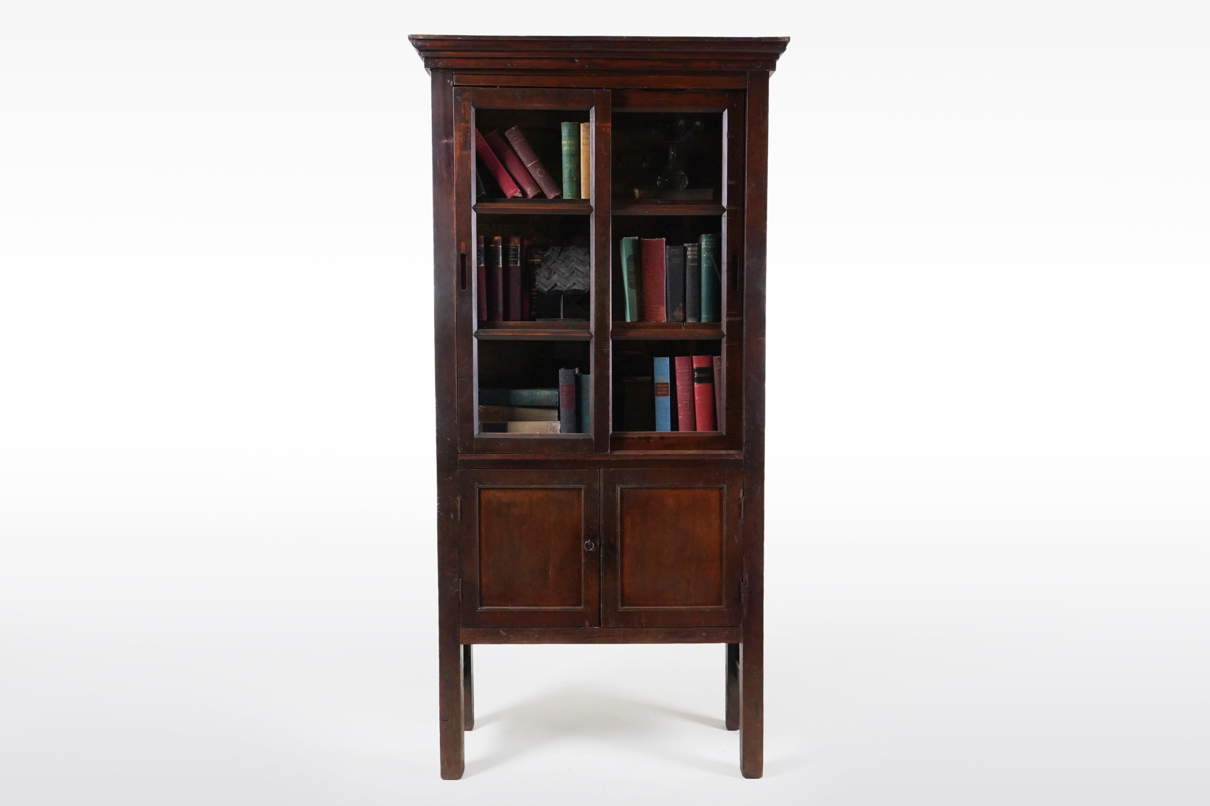A tall British Colonial teak wood storage cabinet with glass doors,c. 1920. This sturdy antique display cabinet features a crown and a tall case with pair of three-panel front doors, set with glass. This piece is made of teak wood, which has
