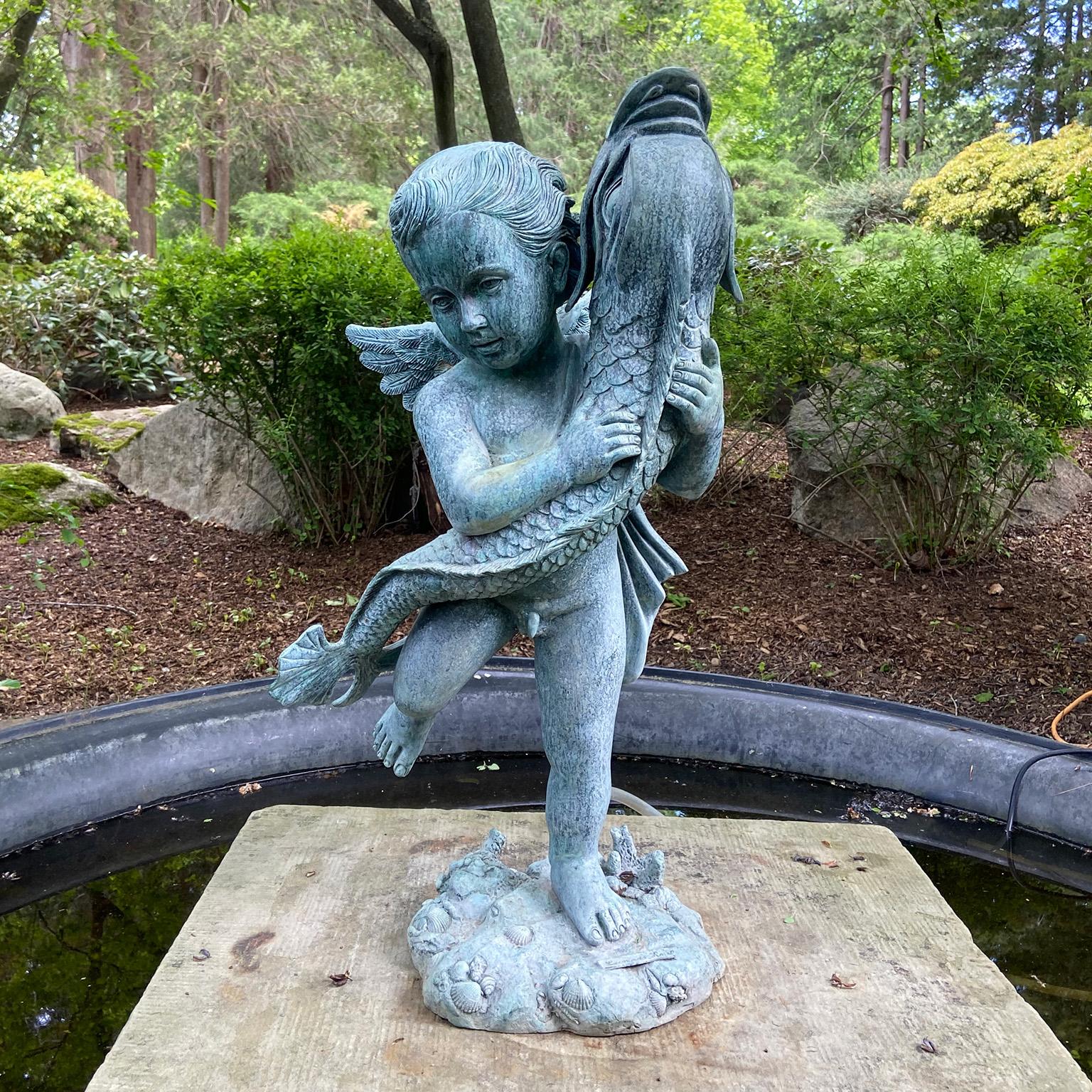 A bronze fountain figure of a winged cherub holding a fish, the figure standing with proper right foot aloft, the right foot standing atop rock-work base with seashells, the fish of the koi variety, the whole in the spirit of Andrea del Verrocchio’s
