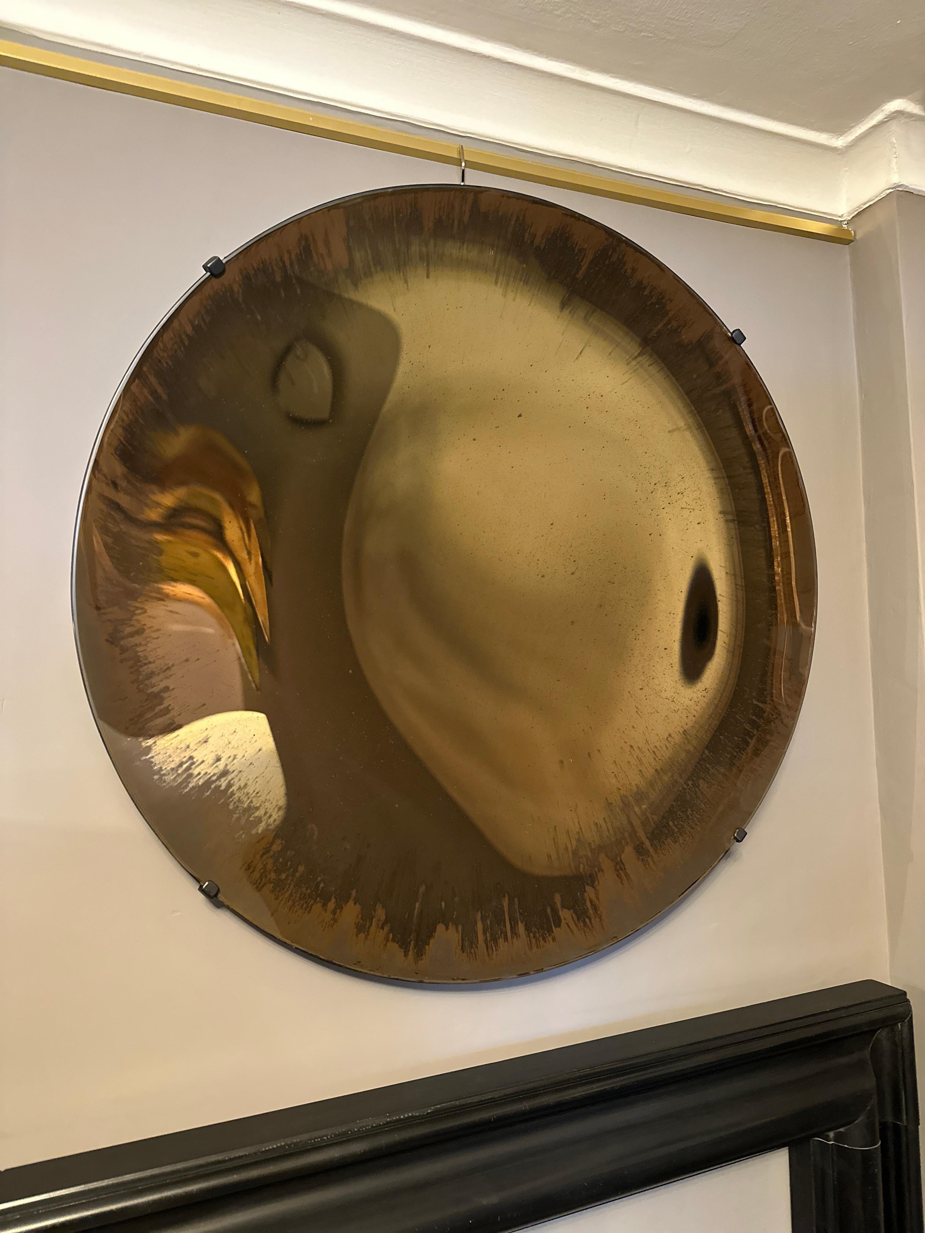 Specchio concavo in vetro color bronzo con finitura anticata. Senza cornice e con supporti da fissare alla parete. Le immagini mostrano i cambiamenti del riflesso. Il colore bronzo intenso è leggermente rovinato sul bordo. Uno specchio contemporaneo