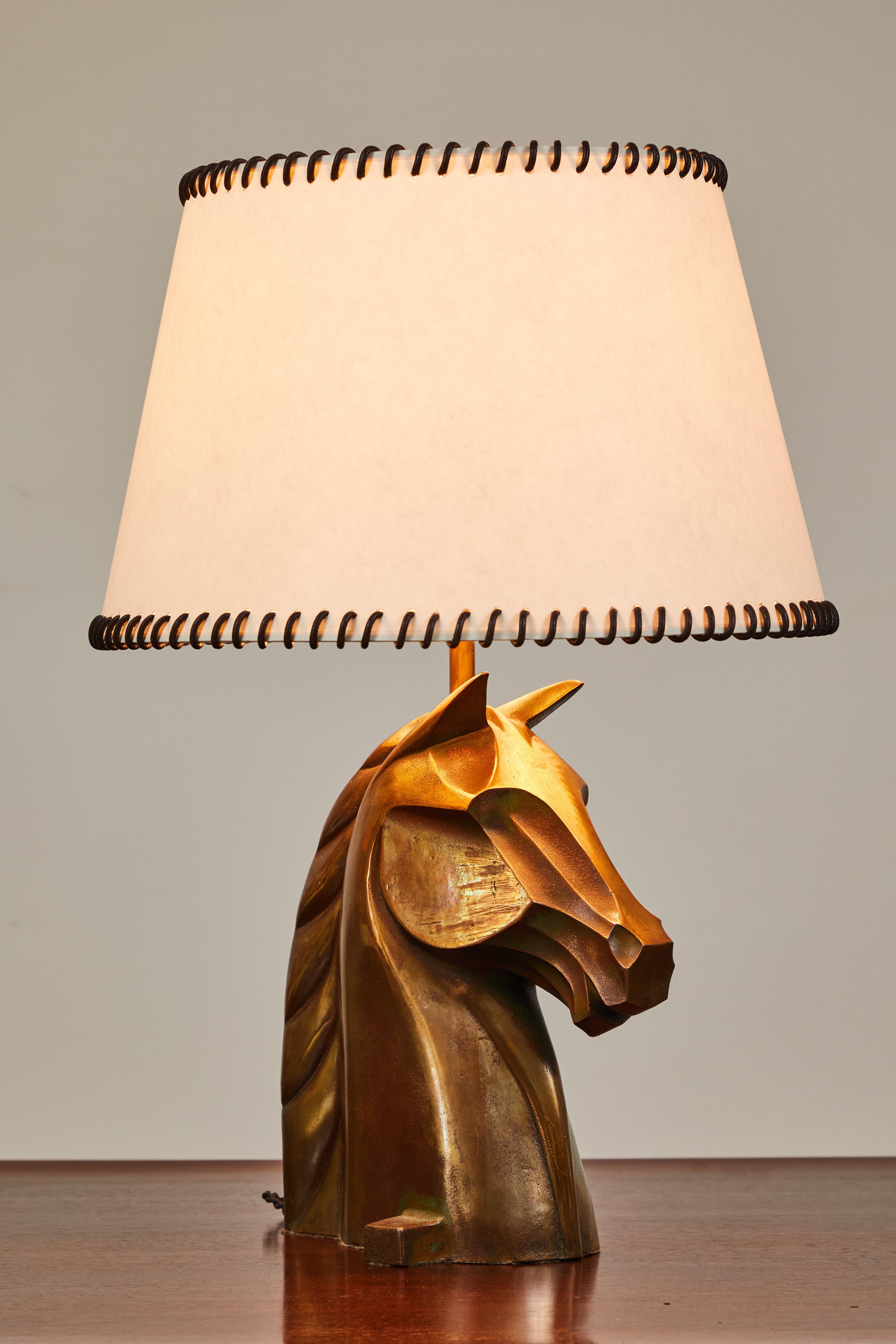 The base of this lamp is a beautiful deco inspired equestrian horse head. Cast in bronze, the horse is illuminated from above when lit. The shade is a soft white paper shade embellished with a dark brown leather whipstitch around both the top and
