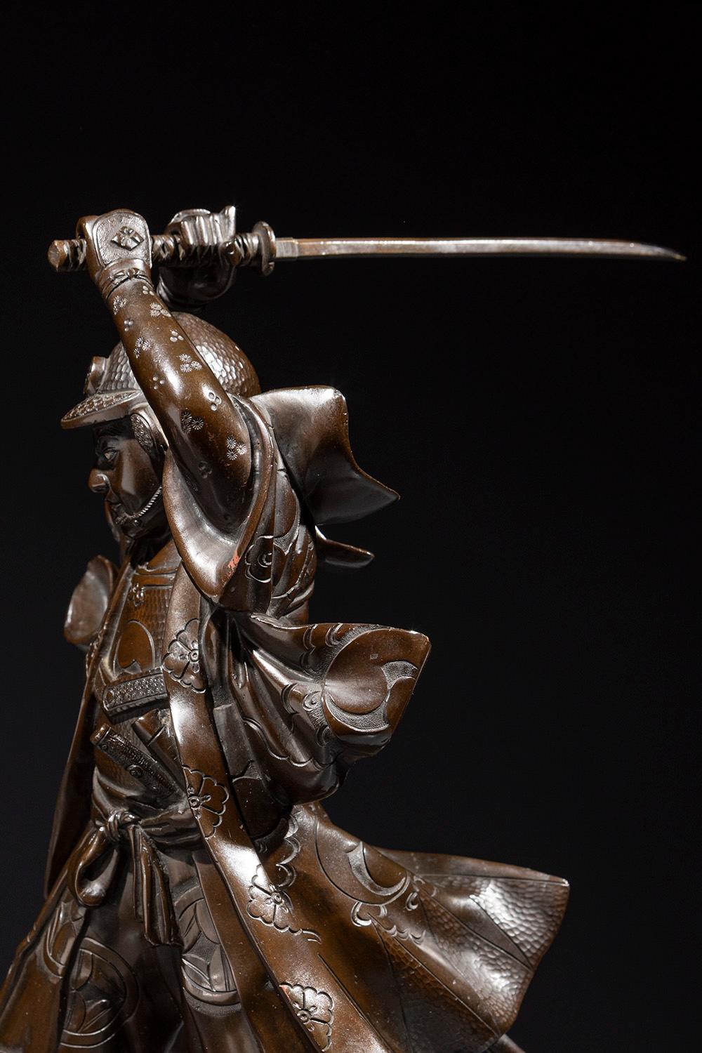 Un okimono in bronzo raffigurante un giovane samurai con la katana sguainata, tenuta con entrambe le mani sopra la testa in posizione di attacco, firmato Yoshimitsu. Lo sguardo e la postura intensi catturano la tensione di un guerriero nel momento