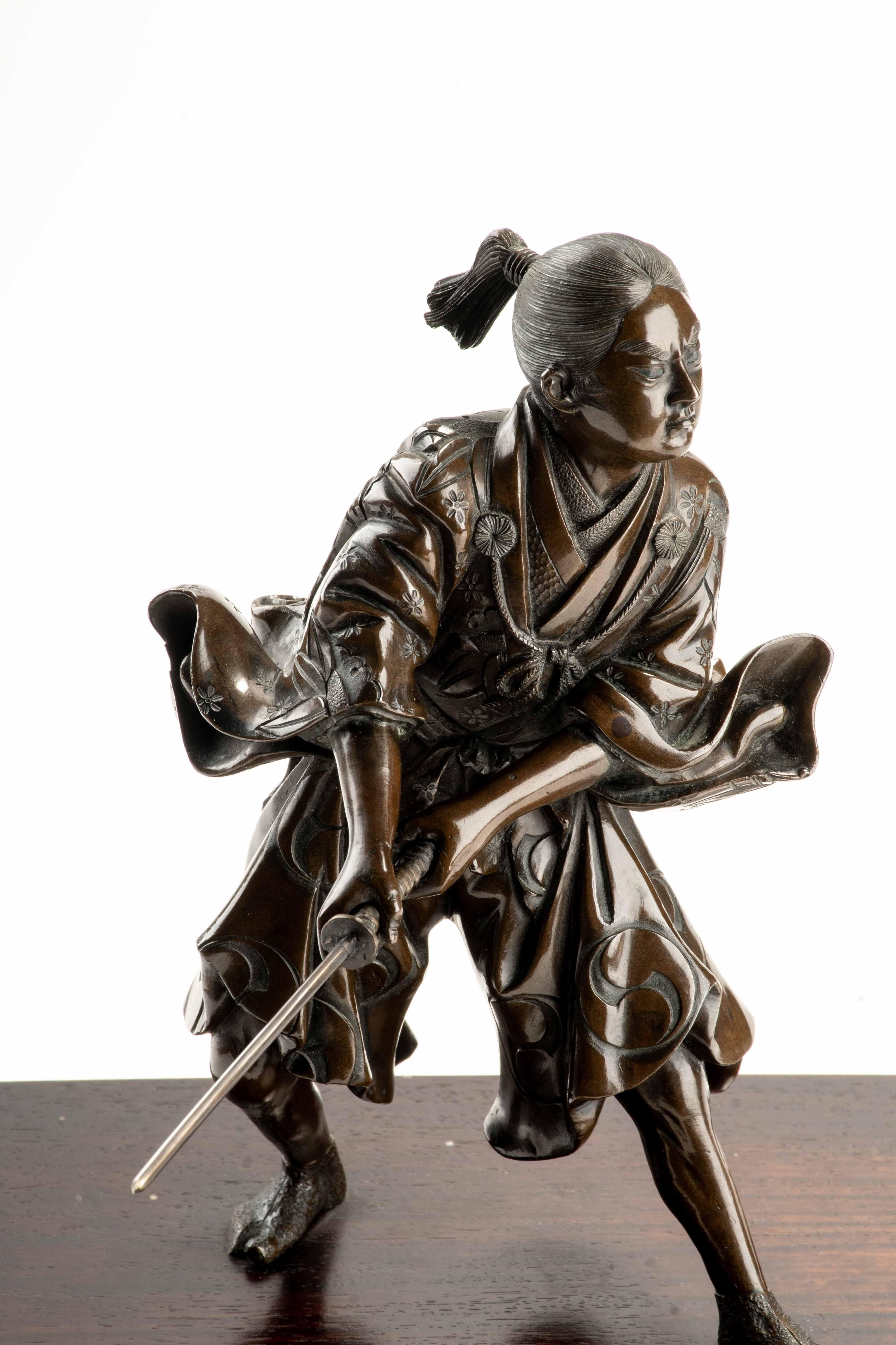 A bronze okimono depicting a young samurai holding a drawn katana with both hands in a guard position.

The kimono is finely engraved with floral motifs, family crests (mon), and wave patterns, with the sleeves animated by movement and wind.

The
