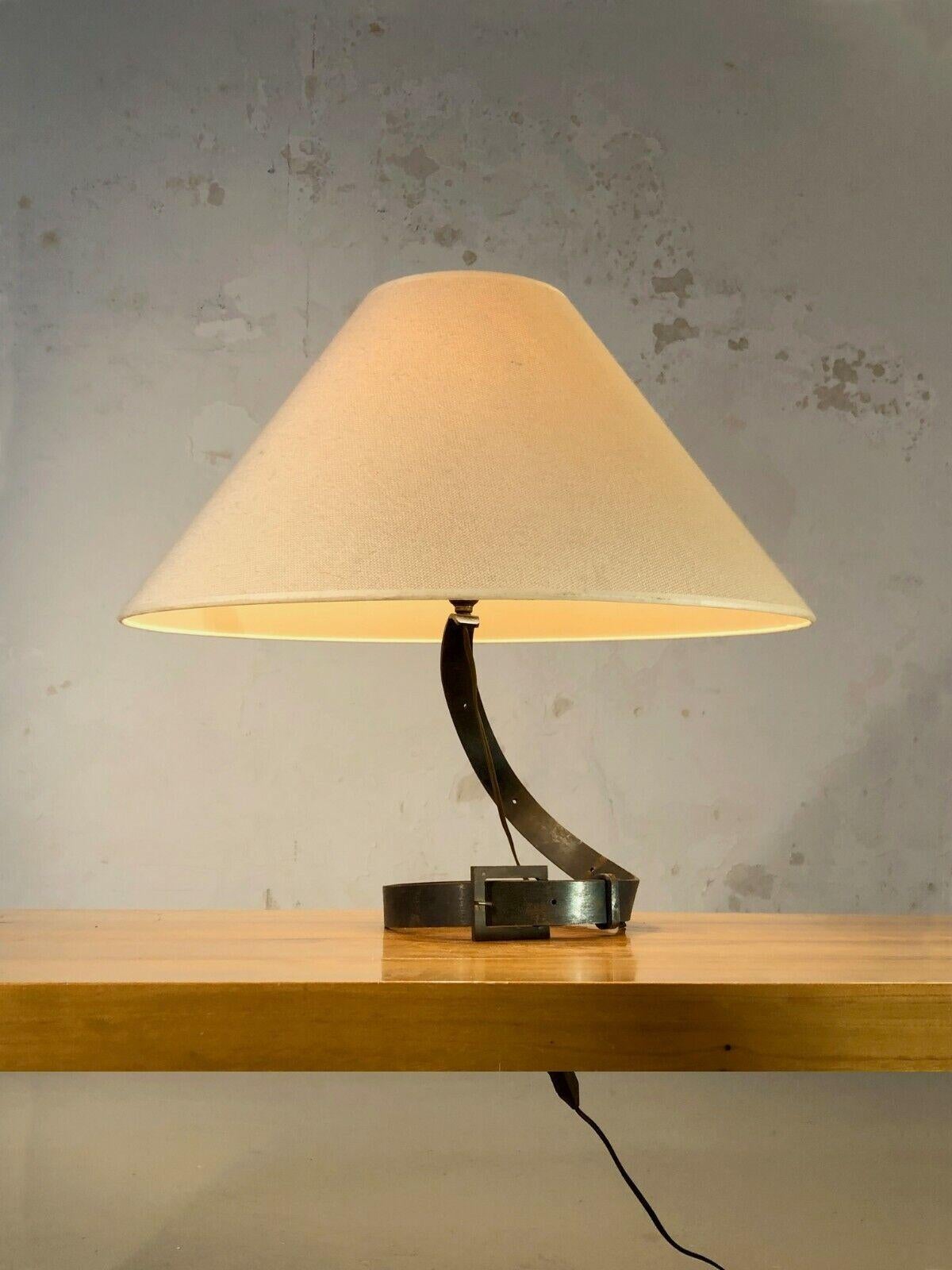 An astonishing and elegant table lamp in metal belt, Modernist, Shabby-Chic, Popular Art, in folded wrought iron decorated with a belt buckle, the whole supporting a socket, to be attributed, France 1960.

SOLD WITHOUT LAMPSHADE OR LAMPSHADE IN