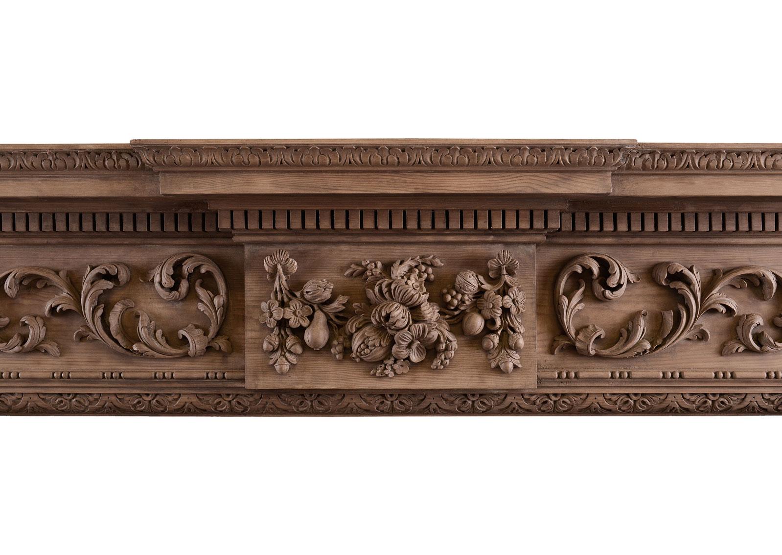 A carved English pine fireplace. The jambs with carved bellflowers surmounted by a cartouche. The frieze with central blocking featuring heavily carved flowers, acorns and leaf work, flanked by scrollwork. The shelf with dentils throughout, and