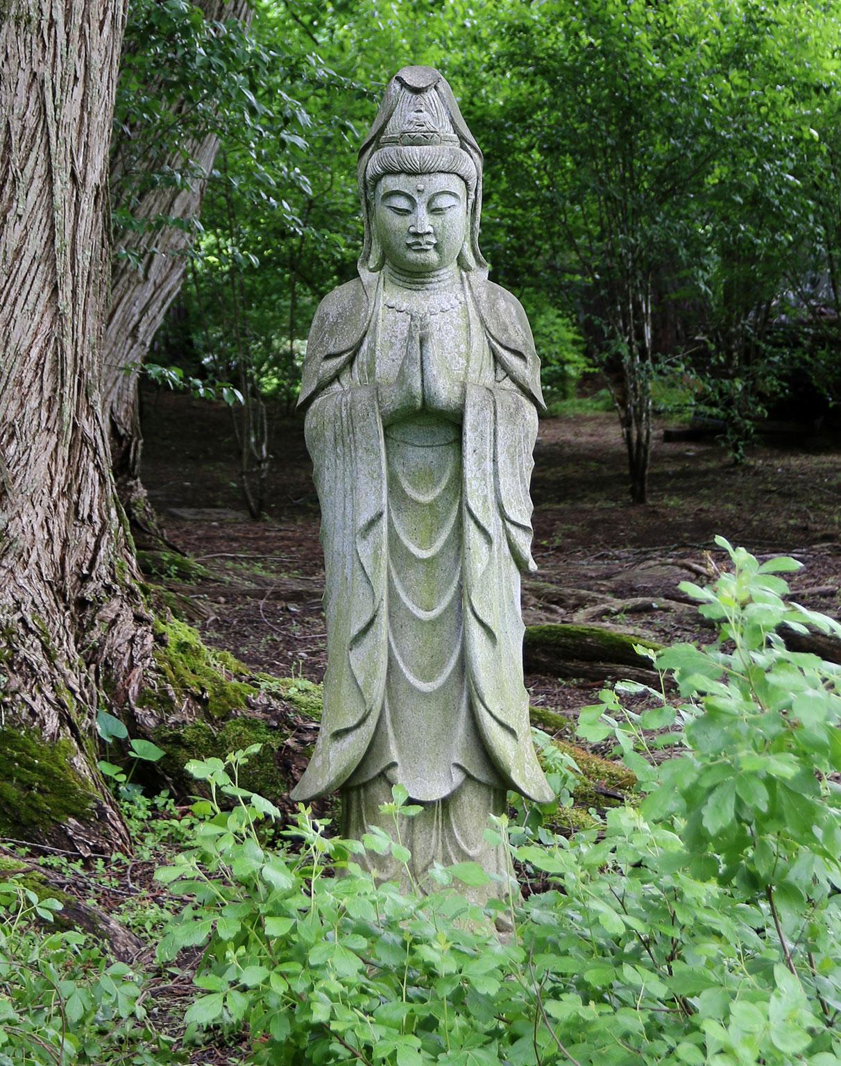 A carved granite figure of Guanyin, one of the most sacred Bodhisattvas in Mahayana Buddhism, shown in traditional robe and headdress and depicted at a moment of solemn repose, her palms joined together in prayer, her head and glance cast