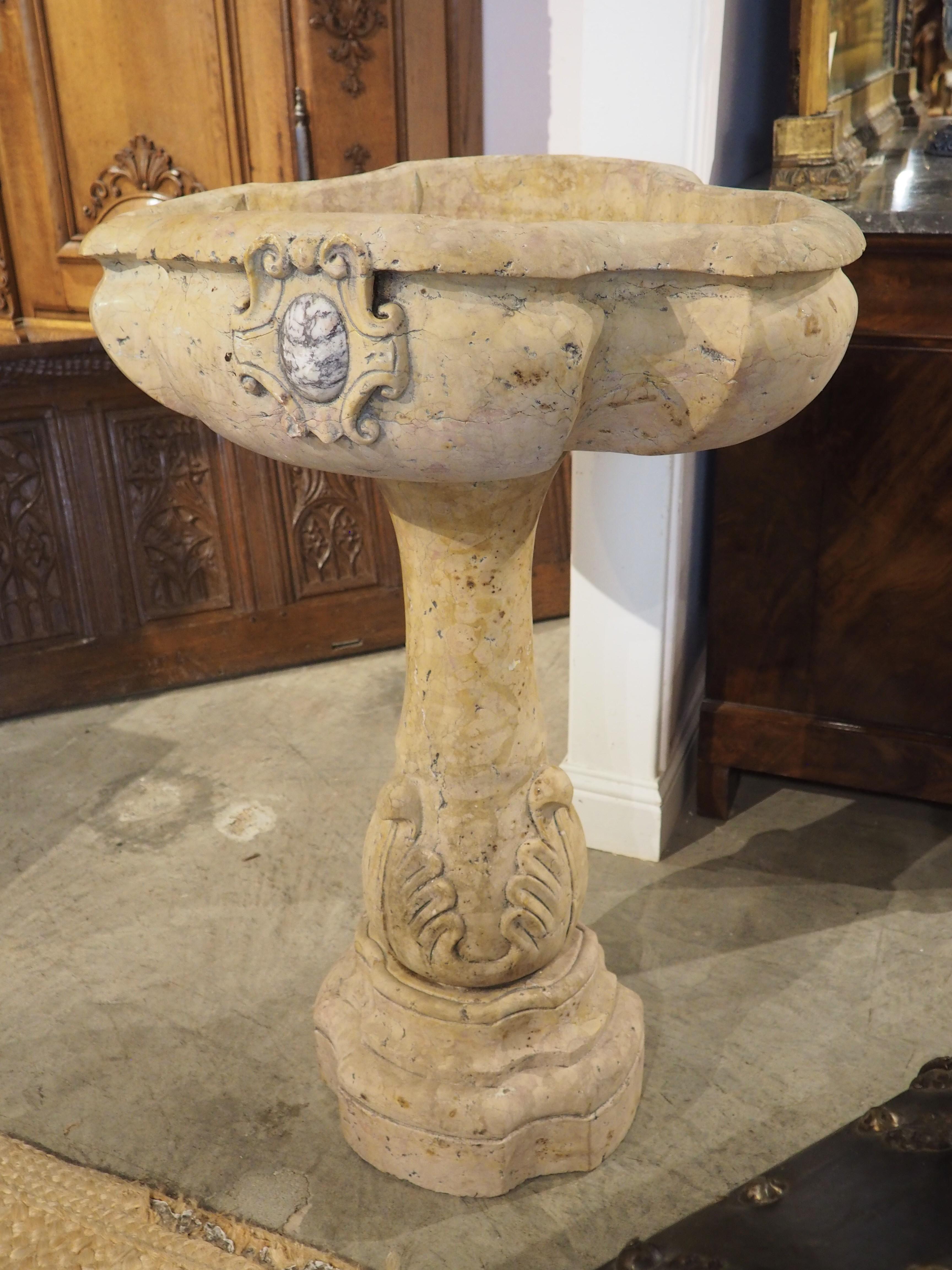 Questa acquasantiera è stata scolpita a mano in un bellissimo e antico marmo Giallo Reale Rosato. Lungo i lati anteriore e posteriore, ci sono dei cartigli che circondano un motivo ovale di uova in marmo bianco e grigio. Il piedistallo sagomato