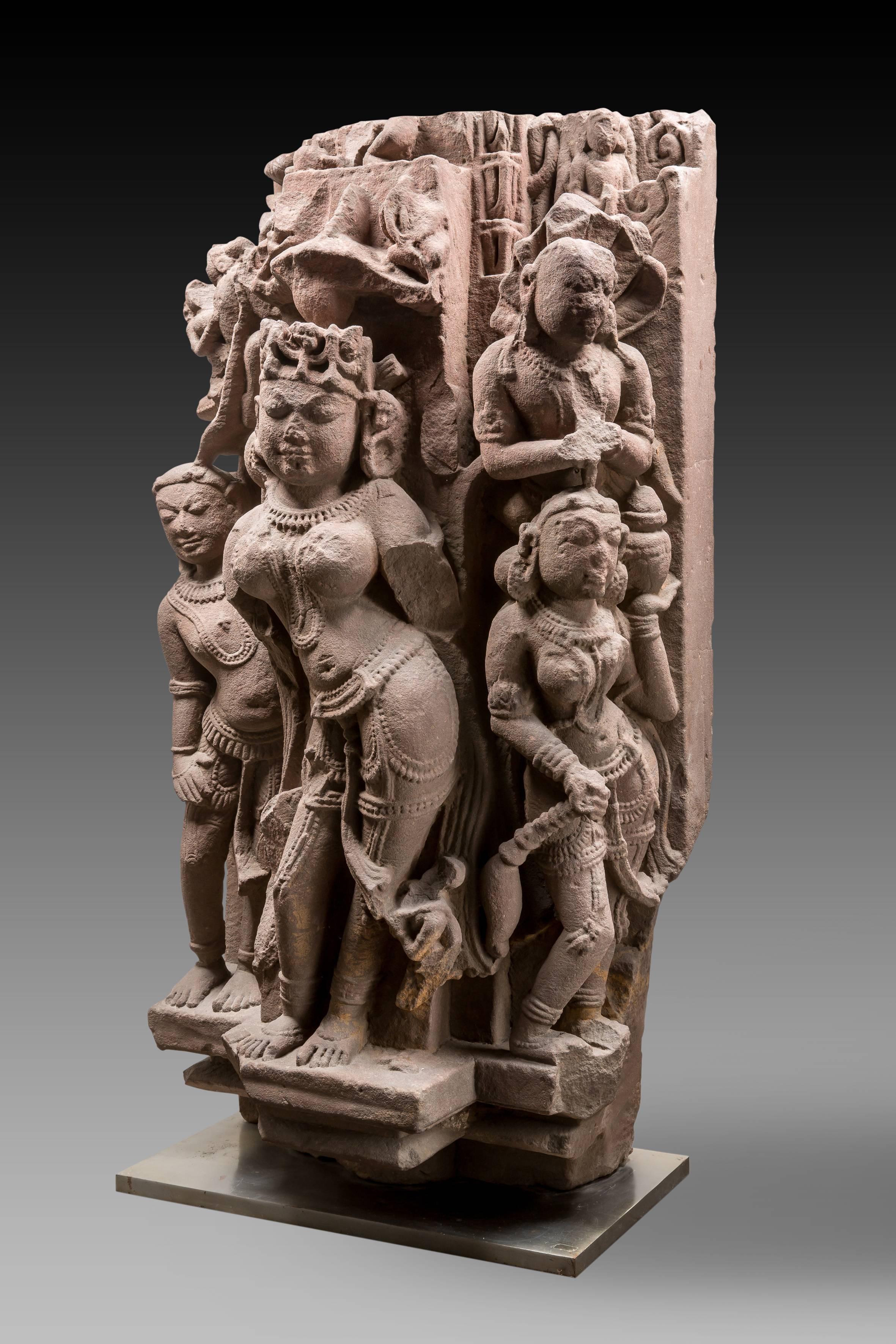The standing female Devi, part of an important architectural relief, is standing in tribangha under a parasol which indicates her status. She is crowned and accompanied by two main attendants, the one on her left is holding a fly chaser and is