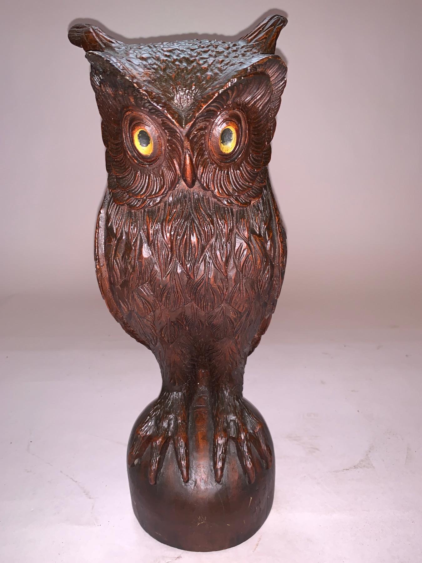 A beautiful majestic carved Owl in one piece in lovely old brown finish with interestingly carved ears and eyes colored in a yellow background with very dark pupils, very large claws and  sitting above a rounded base. On the back is a slightly flat