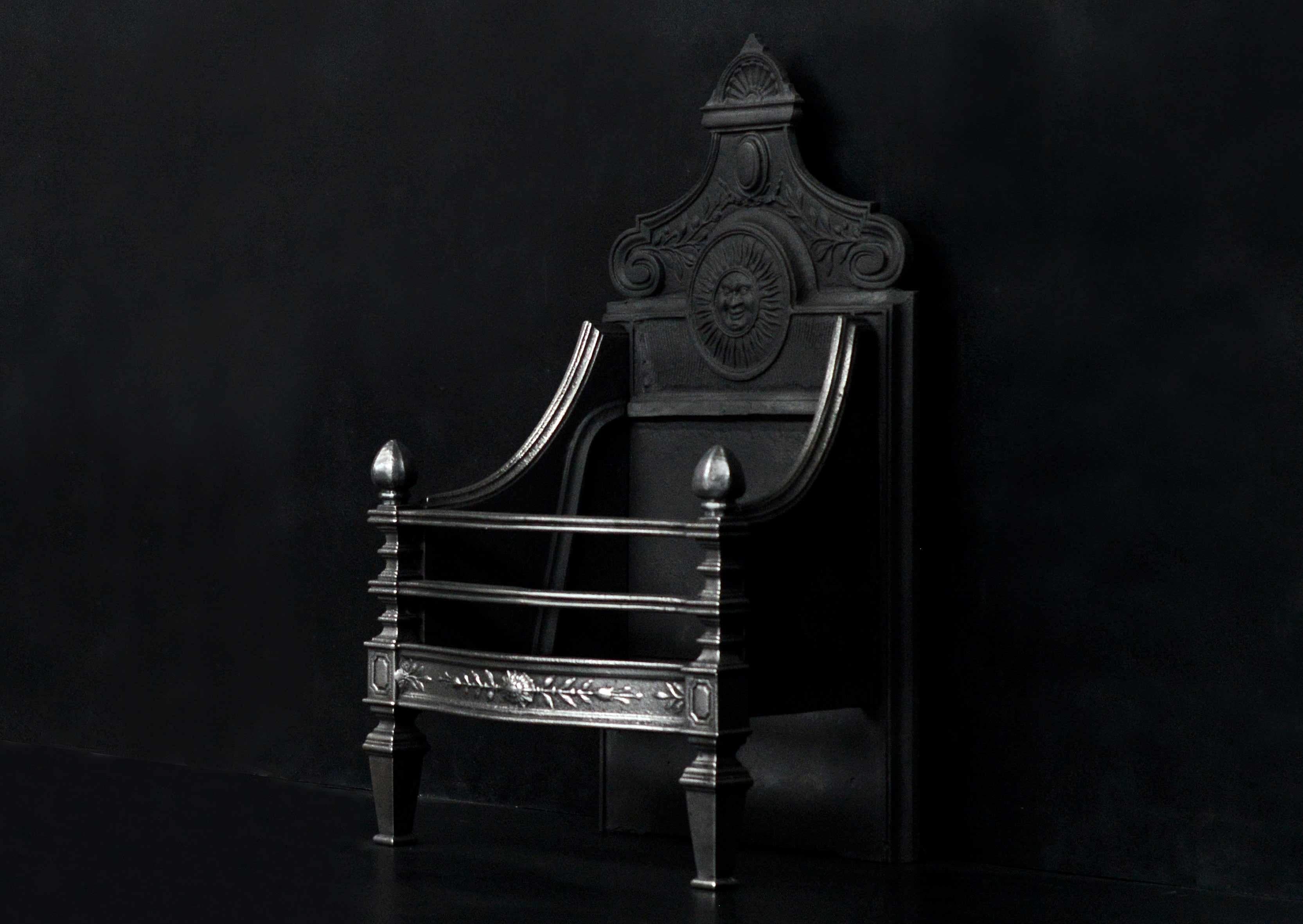 A cast iron English firegrate. The cast fret with decorative leaf work and sunburst paterae to centre, the legs with tapering feet, panelling and hexagonal finials. The cast iron back with sun mask to centre and embellishments above. English, 19th