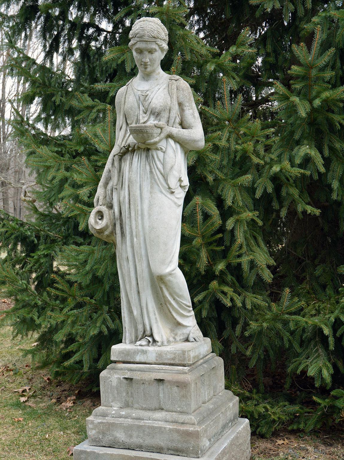 A composition stone figure of Hebe, the goddess of youth and the cupbearer to the gods and goddesses of Mount Olympus, shown in classical dress, holding a cup in her proper left hand and a ewer in her proper right, the figure standing on a stepped