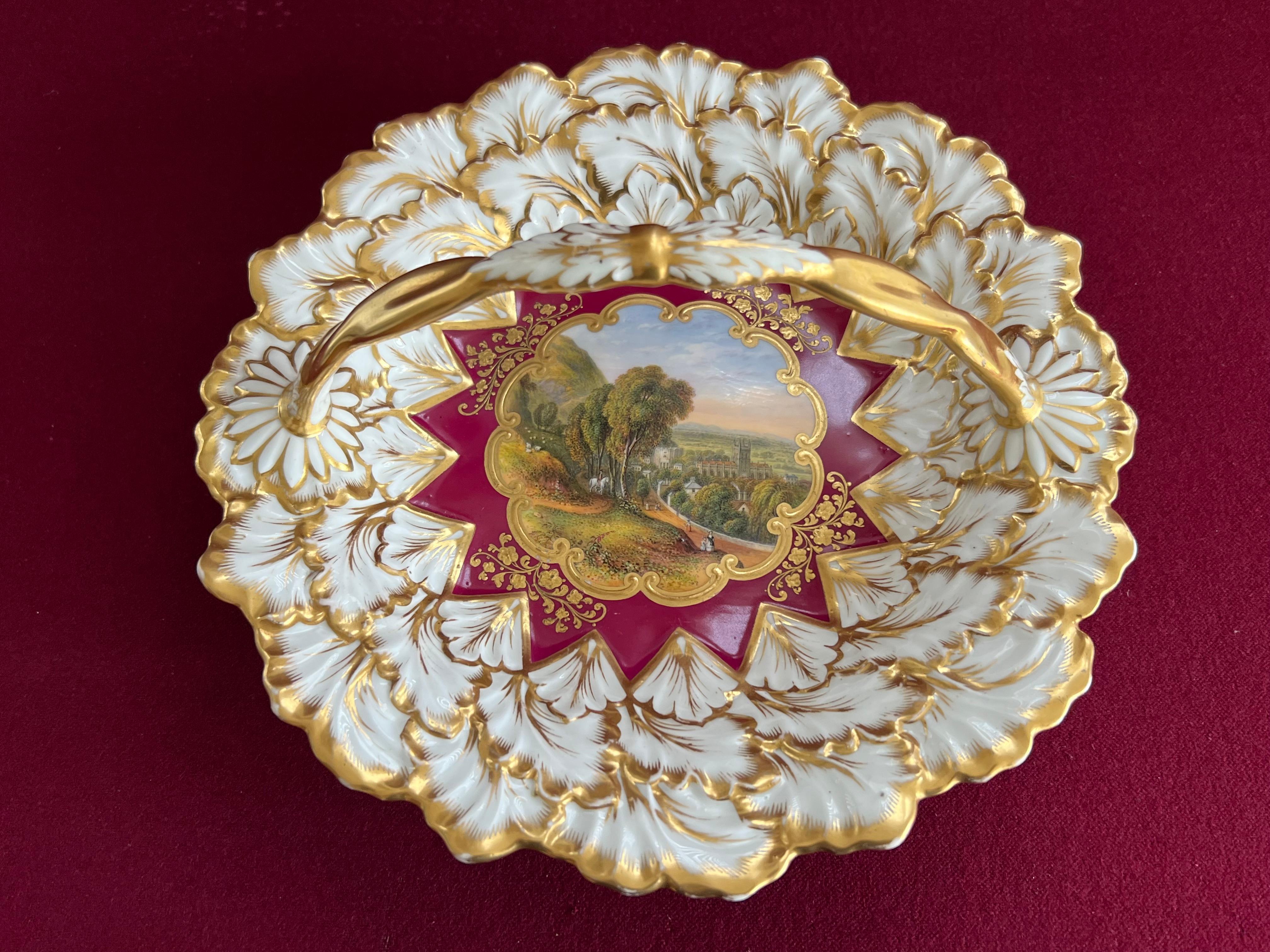 A Chamberlain & Co Worcester porcelain basket c.1845. Of circular form with an overhead handle, moulded with overlapping leaves, the claret red ground reserved with a central cartouche finely painted with a view of Malvern, with raised gilt