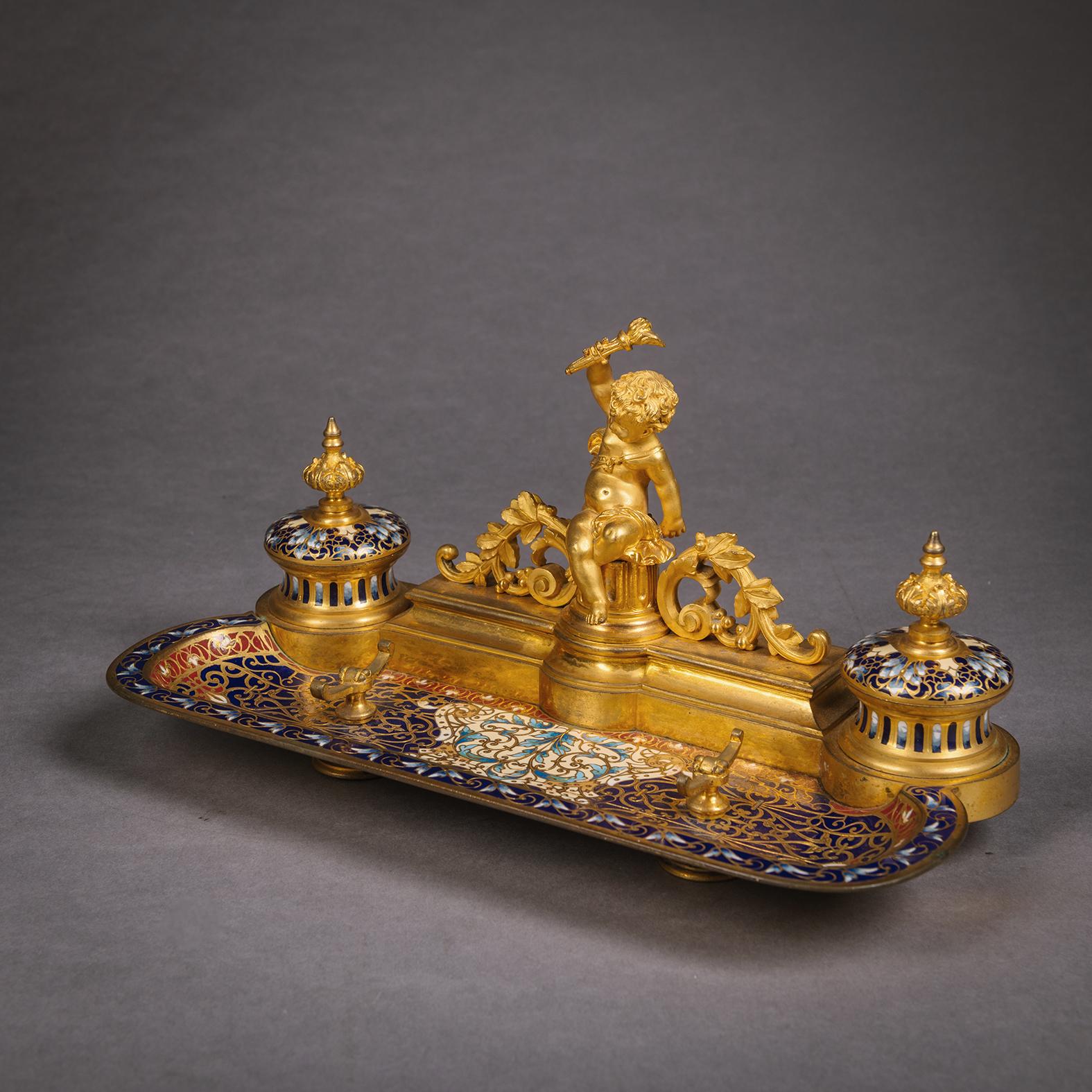 A Champlevé Enamel Three Piece Desk Set, Attributed to Barbedienne. 

Comprising a gilt-bronze pen tray and a pair of candleholders all finely decorated in Champlevé enamel.  The pen tray with two hinged lidded ink wells with a stepped gallery