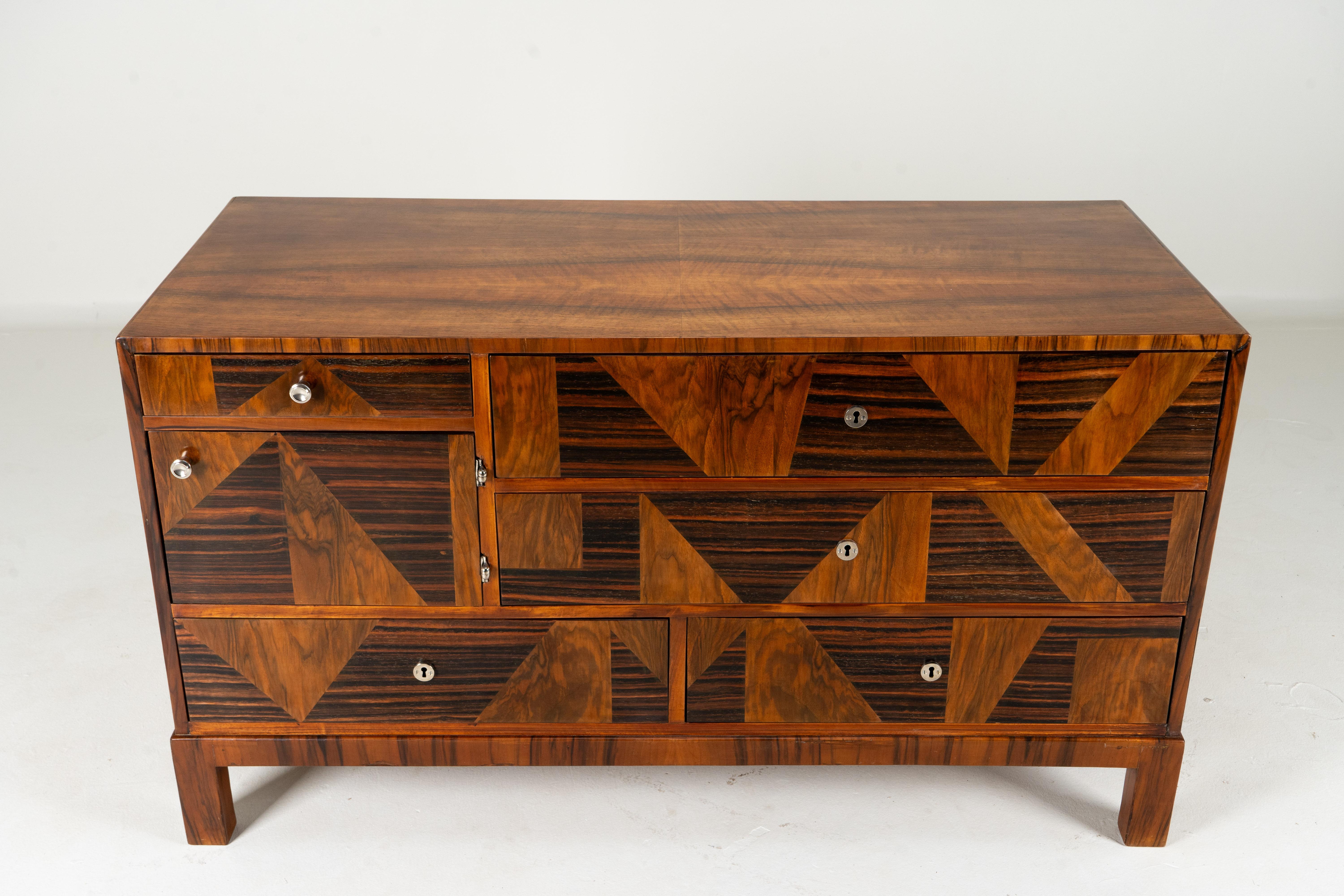 This Hungarian chest of drawers dates to the late 1940's or early 1950's and features Mid-Century Modern styling with an Art Deco twist. The piece is crafted with a walnut case and macassar veneer inserts to the door and drawer fronts. The piece has