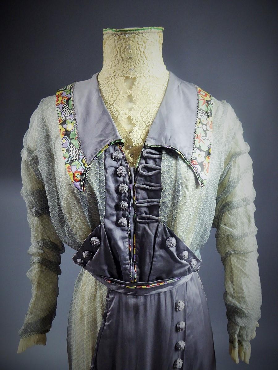 Vers 1905/1910
Angleterre 
Une belle robe de thé d'une dame anglaise datant du début du vingtième siècle. Satin gris et mousseline imprimés de motifs circulaires. Effet d'un tablier et d'une bavette agrémentés de boutons dans le même nœud brodé.