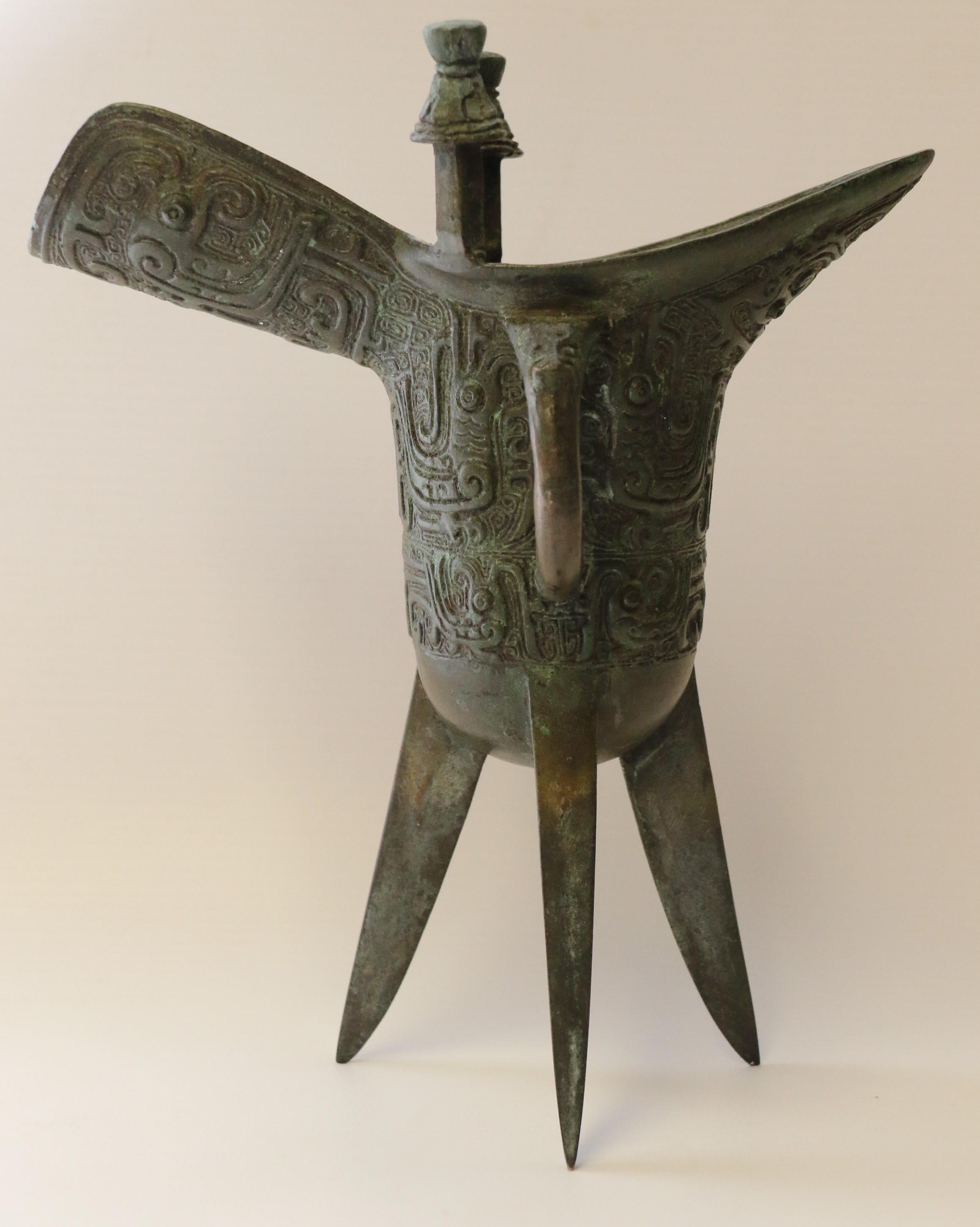 This large archaic style bronze jue is an ancient Chinese wine vessel that originally dates to 1600-1046 BC was used to heat and serve wine in ceremonial purposes. The tripod long legs would sit over hot embers to heat the wine and the two