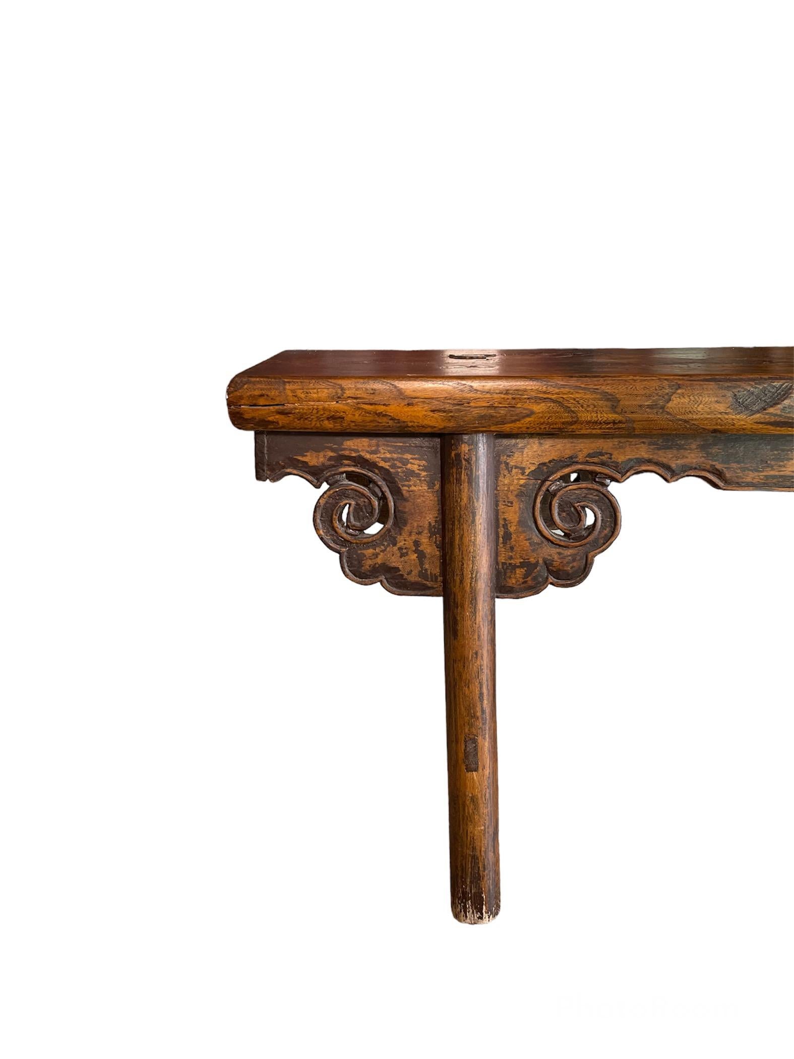 This is a Chinese countryside hand crafted wood bench. It depicts a small rectangular top with two pair of cylindrical legs joined by stretchers. Below the top, there is a skirt with scalloped border that surrounds the table and ends with scrolls at