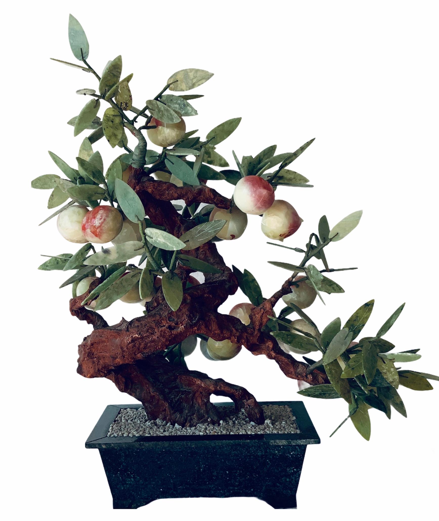 This is a peach tree bonsai. The peaches are made of coral yellow green colored hardstones and the leaves are made of nephrite jade. The tree is standing in a dark green marble rectangular planter that is full of white pebbles. There are some
