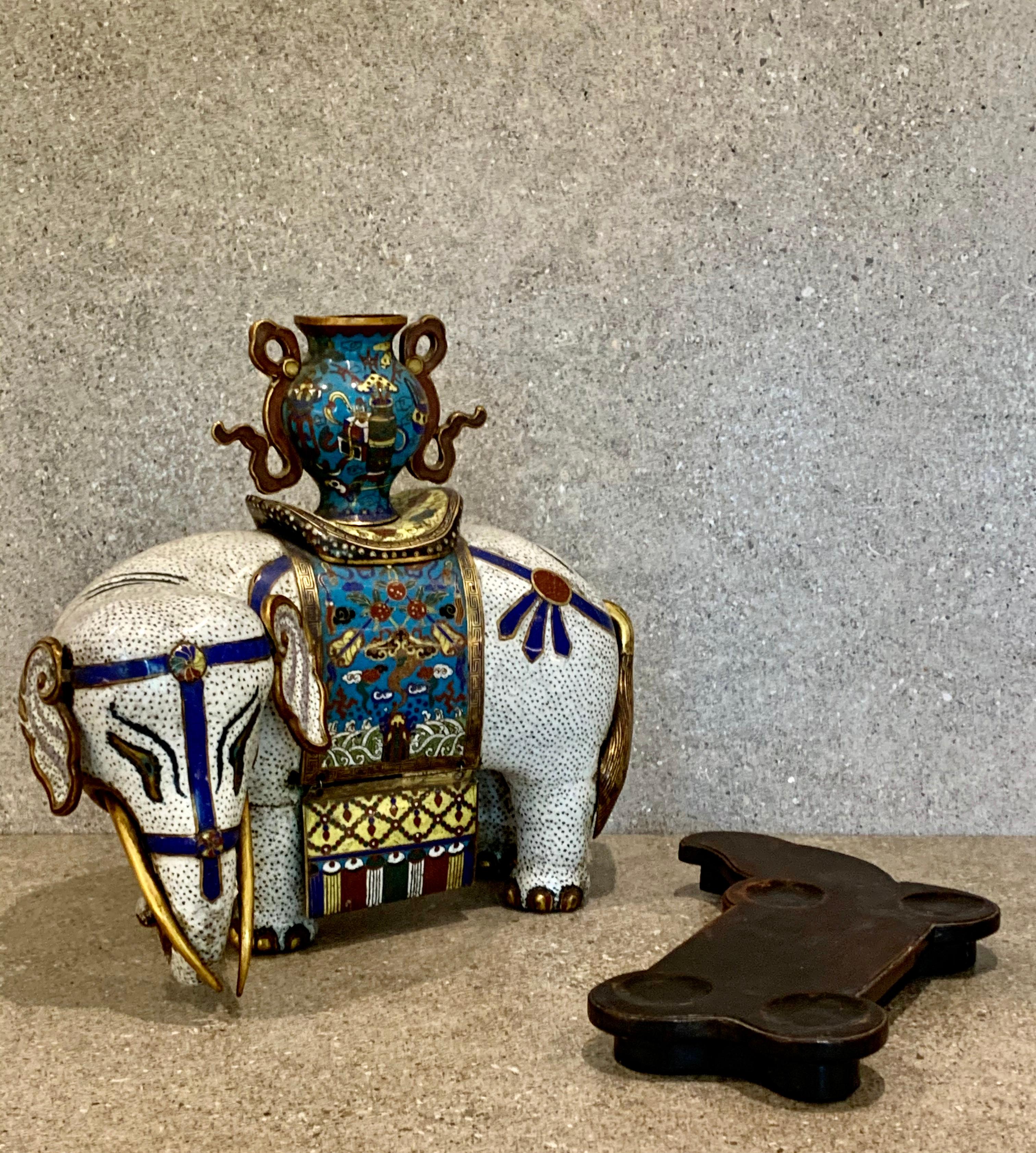 The white elephant stands turning its head to sinister, its saddle blanket decorated with fruits and precious objects over crashing waves, with a vase decorated with antiques upon the animal's back, the ears removable, together with a wood stand,