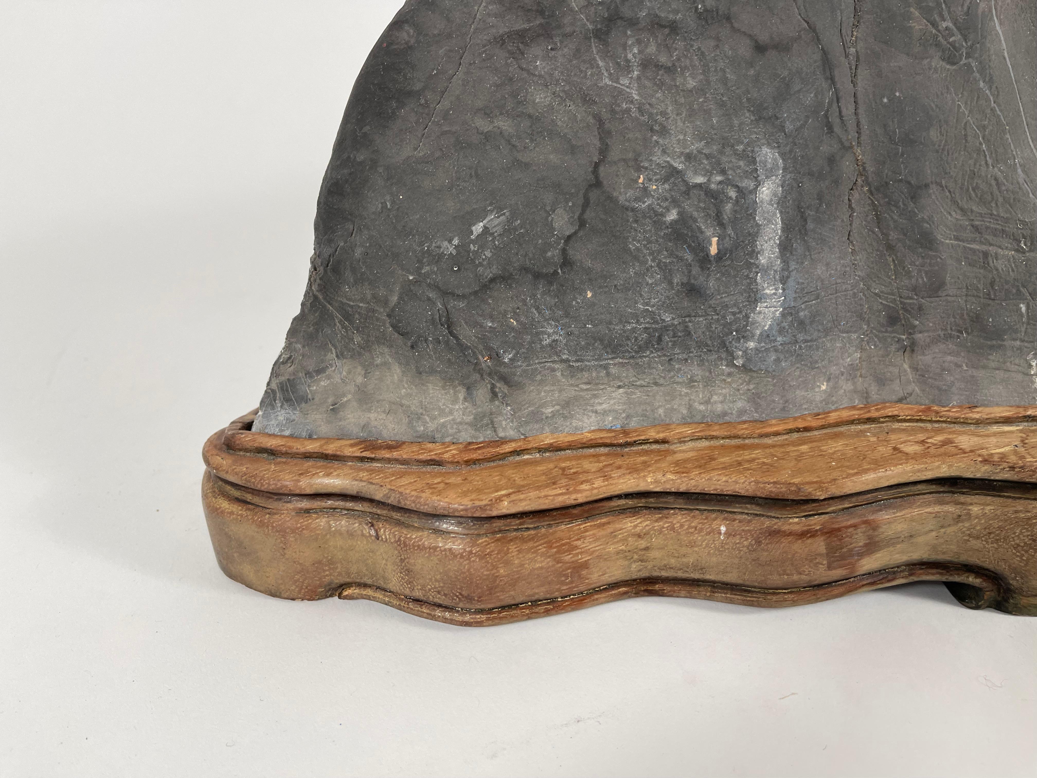 A Chinese or Japanese scholar's rock resembling a mountain range, this specimen with white veins, crevices and smooth peaks, is mounted on a conforming carved wood base. Known as Gongshi in Chinese, and Suiseki in Japanese, scholar's rocks are