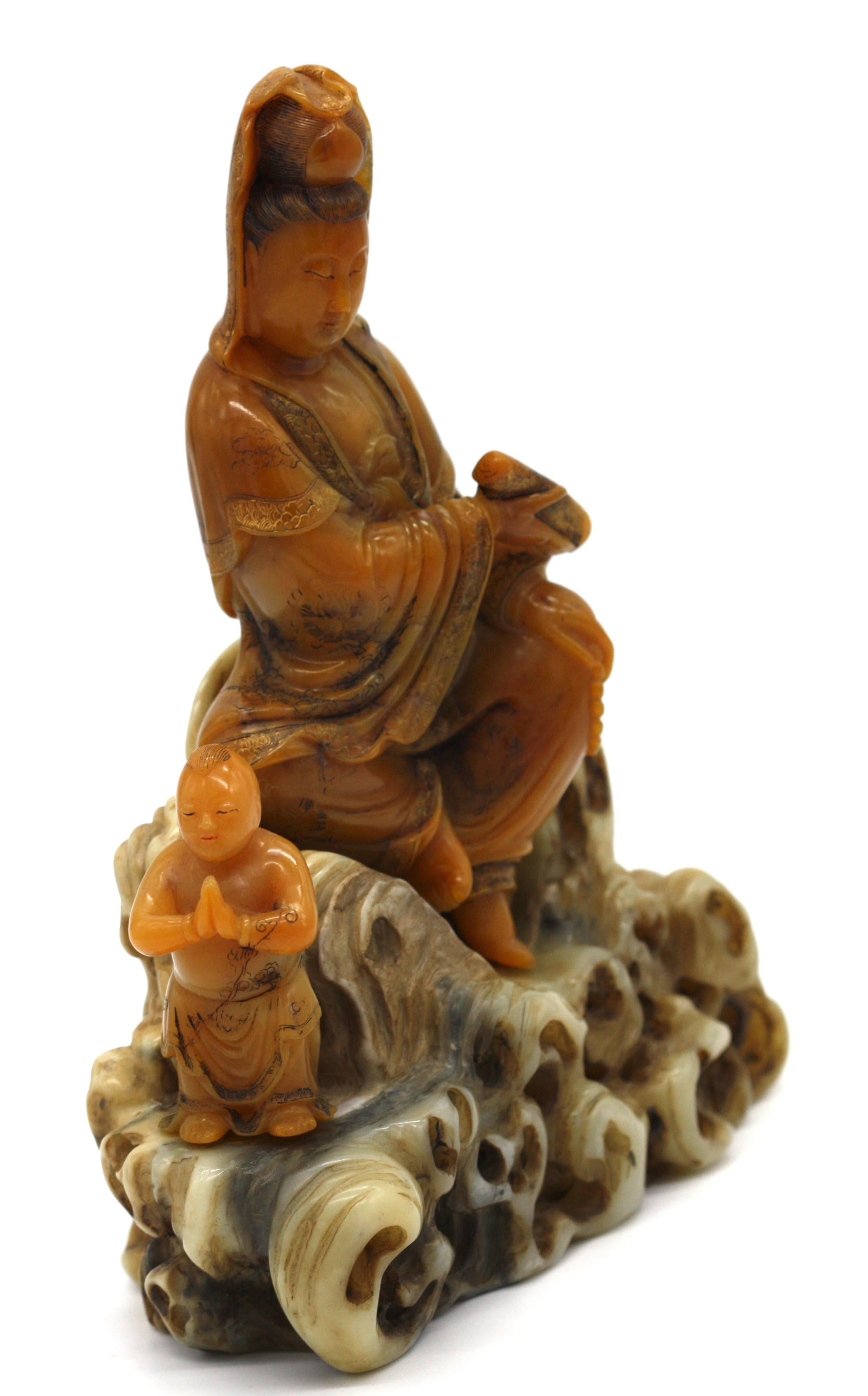 Figurine d'Avalokiteshvara en stéatite chinoise
sculptée assise à côté d'un garçon debout, vêtue d'une robe ample tombant en cascade et délicatement gravée de rinceaux, tenant un rouleau dans une main, son visage à l'expression bienveillante