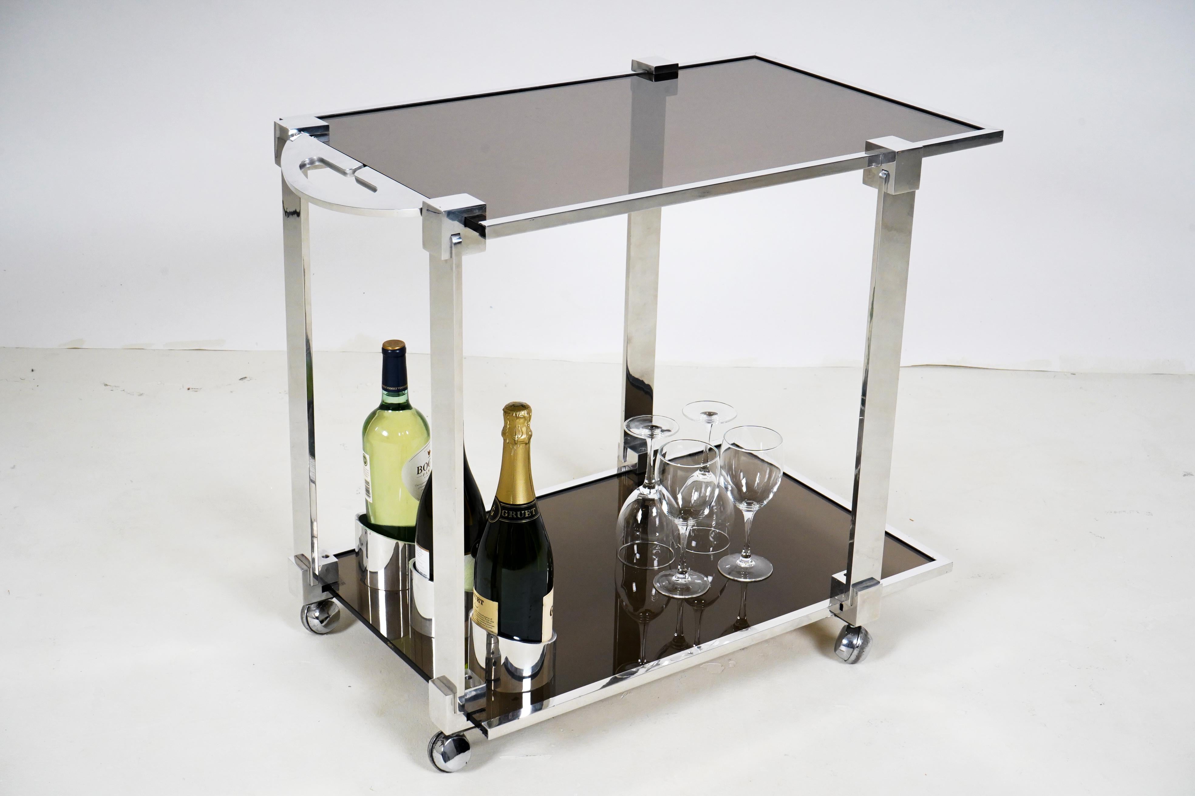 This 1970s chic chrome bar cart has two levels for storage and serving area and 3 bottle holders, also chromed. The top and bottom shelf are fitted with smoked glass, producing a piece that reproduces some of the glamor and style of interiors by