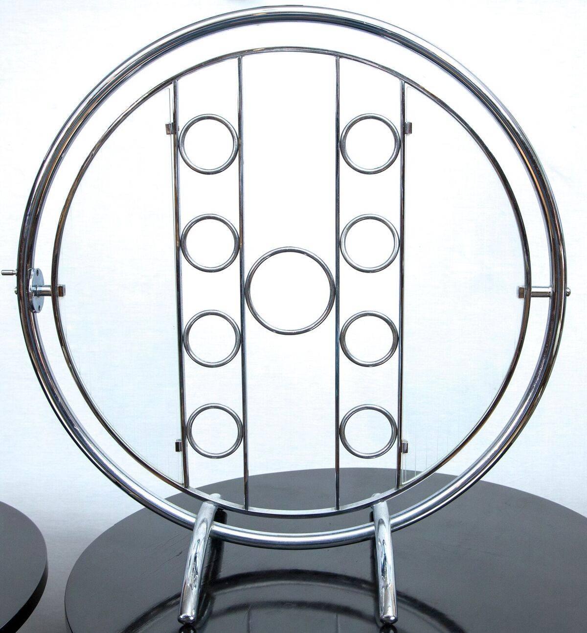 An Art Deco, machine age cocktail table. The table features a round tubular chrome metal framed top, which is held in a swivel frame that allows it to be turned vertically when not in use. The tabletop suspends two half-circle etched glass shelves