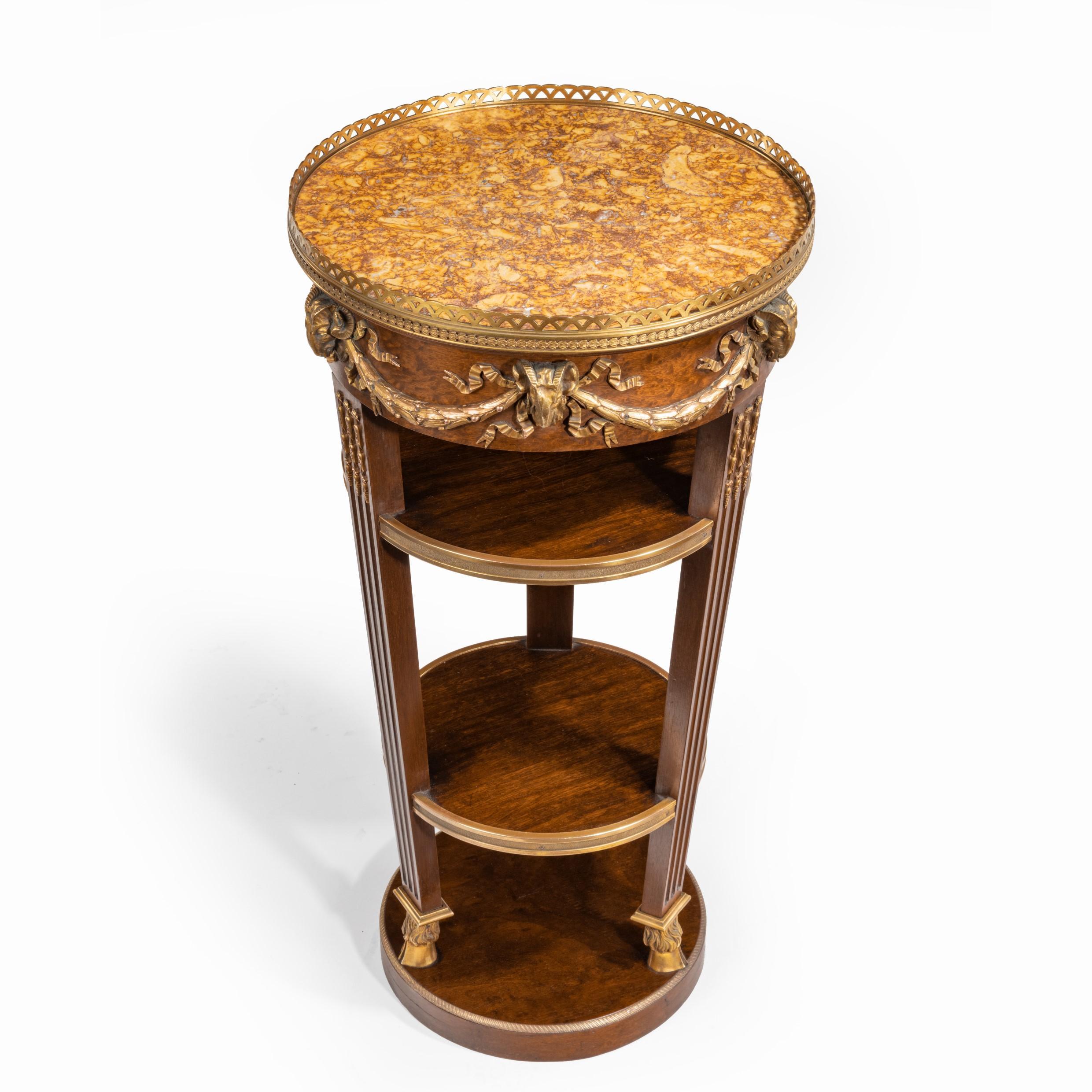 A circular mahogany guéridon in manner of Zwiener, of tapering cylindrical form with two shelves, the circular top with a yellow marble top and ormolu gallery, applied ormolu edges and mounts of rams’ heads strung with swags and raised on three