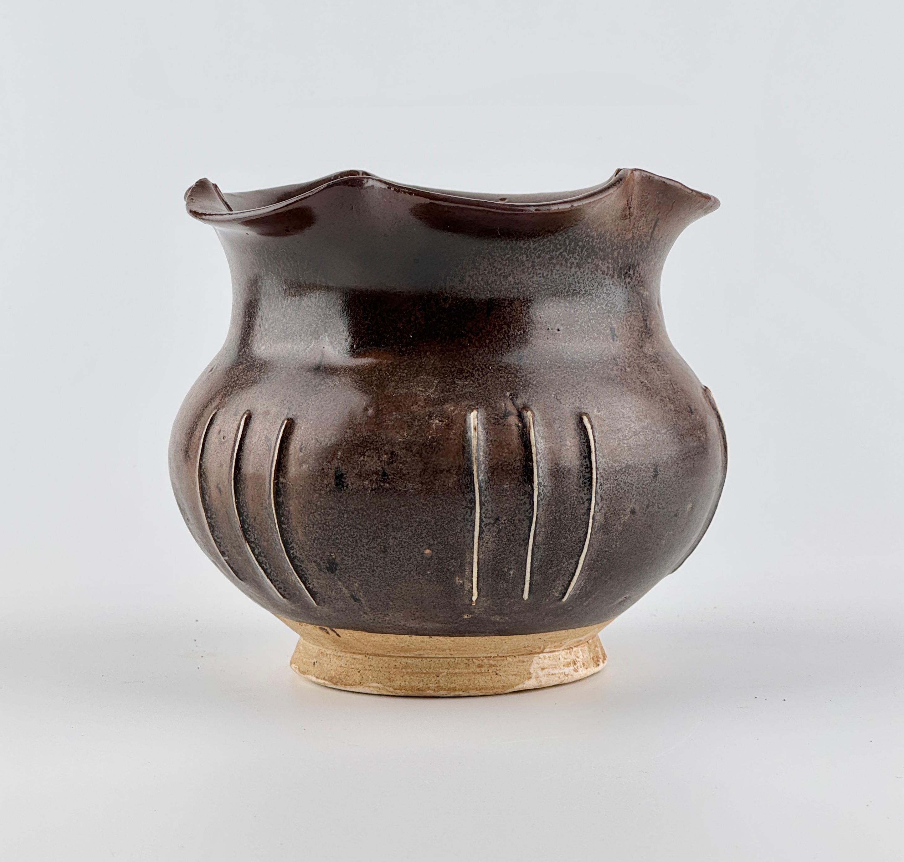 The jar is covered with a deep, warm blackish-brown glaze. The glaze is smooth and even, with a soft sheen.

The foliate rim has a gentle, wave-like design that gives the jar a natural and organic feel. The body features vertical incised lines that