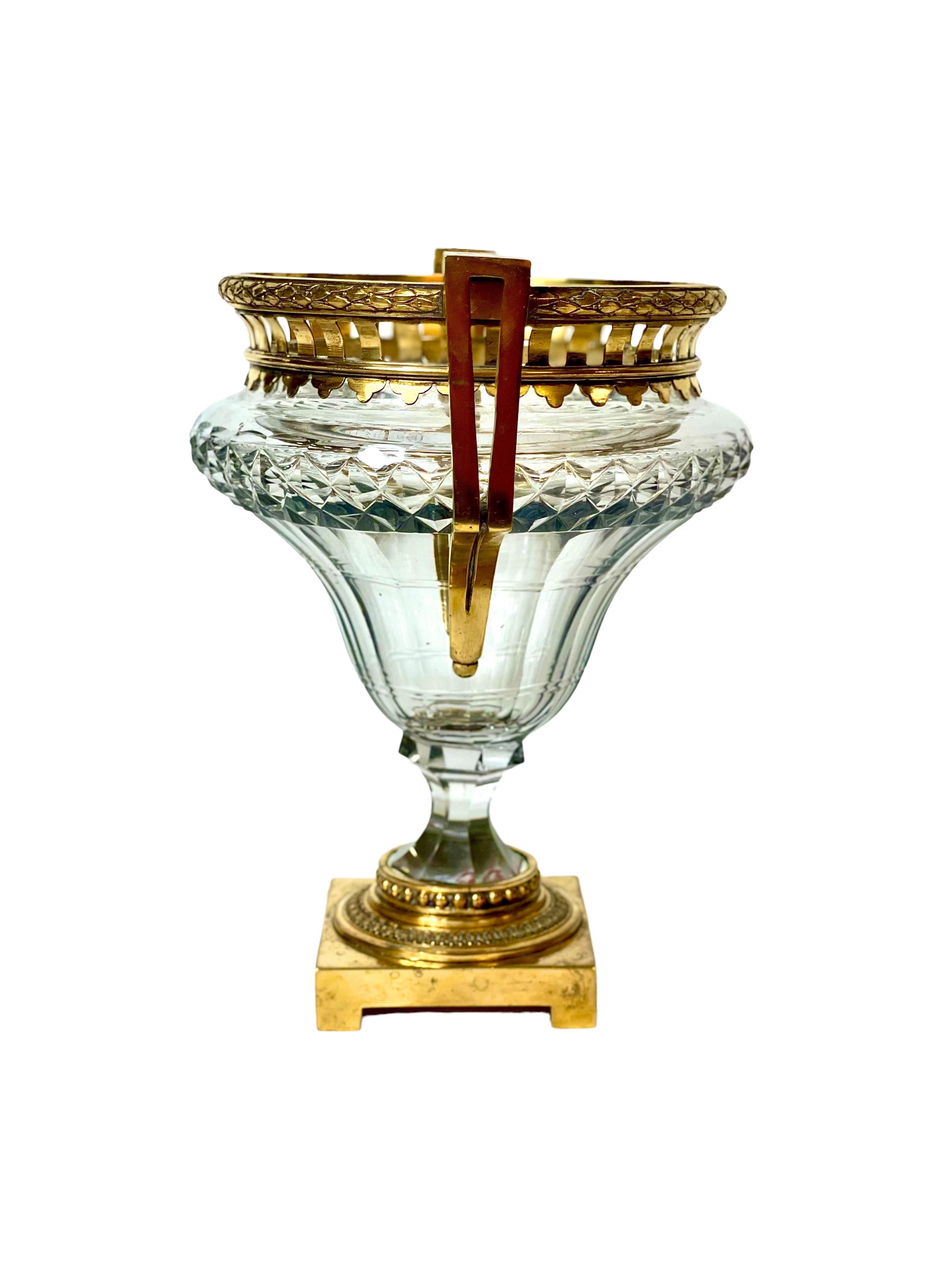 A classic and opulent crystal and gilt bronze Louis XVI-style vase, its goblet-shaped heavy crystal bowl diamond cut and fluted to refract the light. Luxuriously embellished with a brass frame and striking, square-cornered handles, the rim and