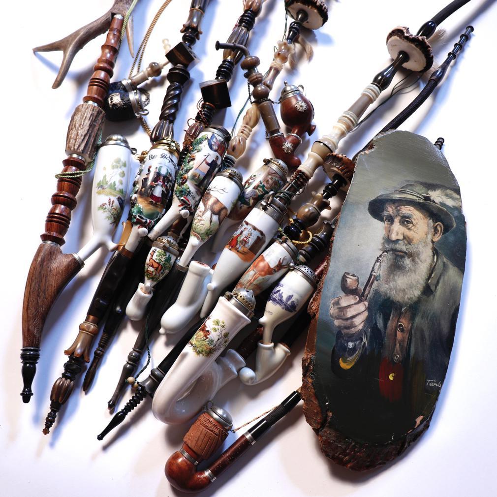 A collection of 13 traditional German porcelain tobacco pipes and a painting. A compositional long stem pipe having a porcelain bowl called the abguss, a tobacco bowl oblong in shape and open at the top, capped by a hinged metal lid, narrowing down