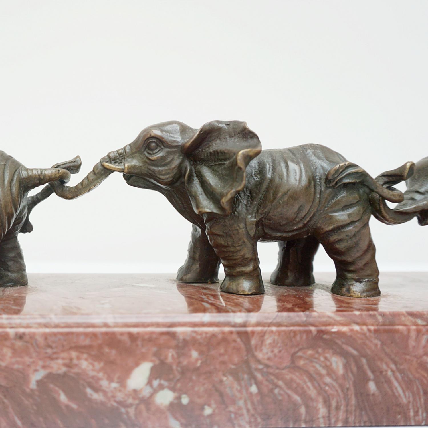 A Contemporary bronze sculpture of a line of playful elephants, connected by their trunks and tails. The elephants growing in size with the largest Elephant at the head of the herd. Set on a marble base. Added signature 'E. Barillot' to marble base.