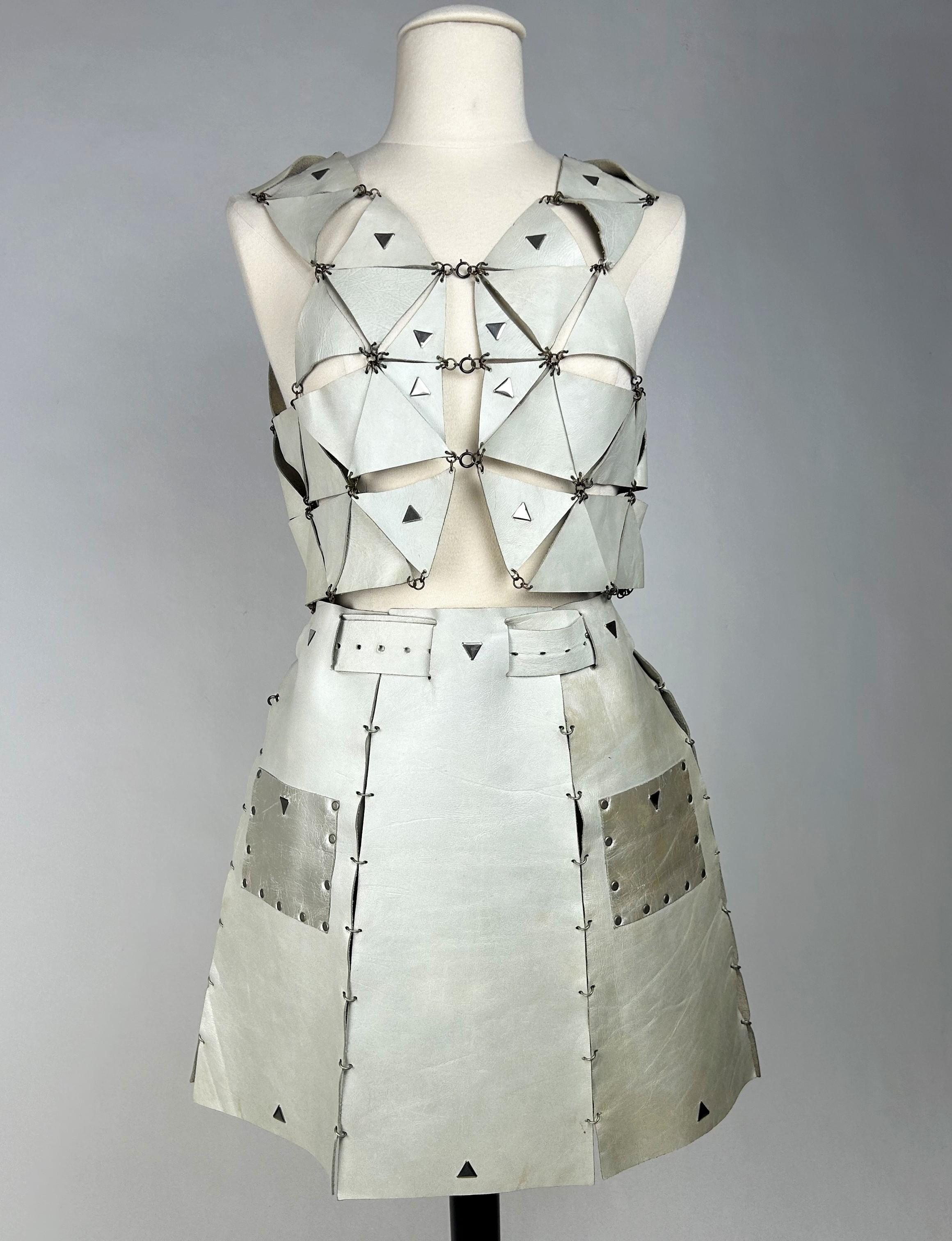 Circa 1967

Paris

Outfit in leather by Paco Rabanne, mini-skirt and open bolero with triangular ivory leather cut-outs held in place by metal rings. Skirt with flared panels appliqué with silvered pockets and brushed steel triangular studs, with an