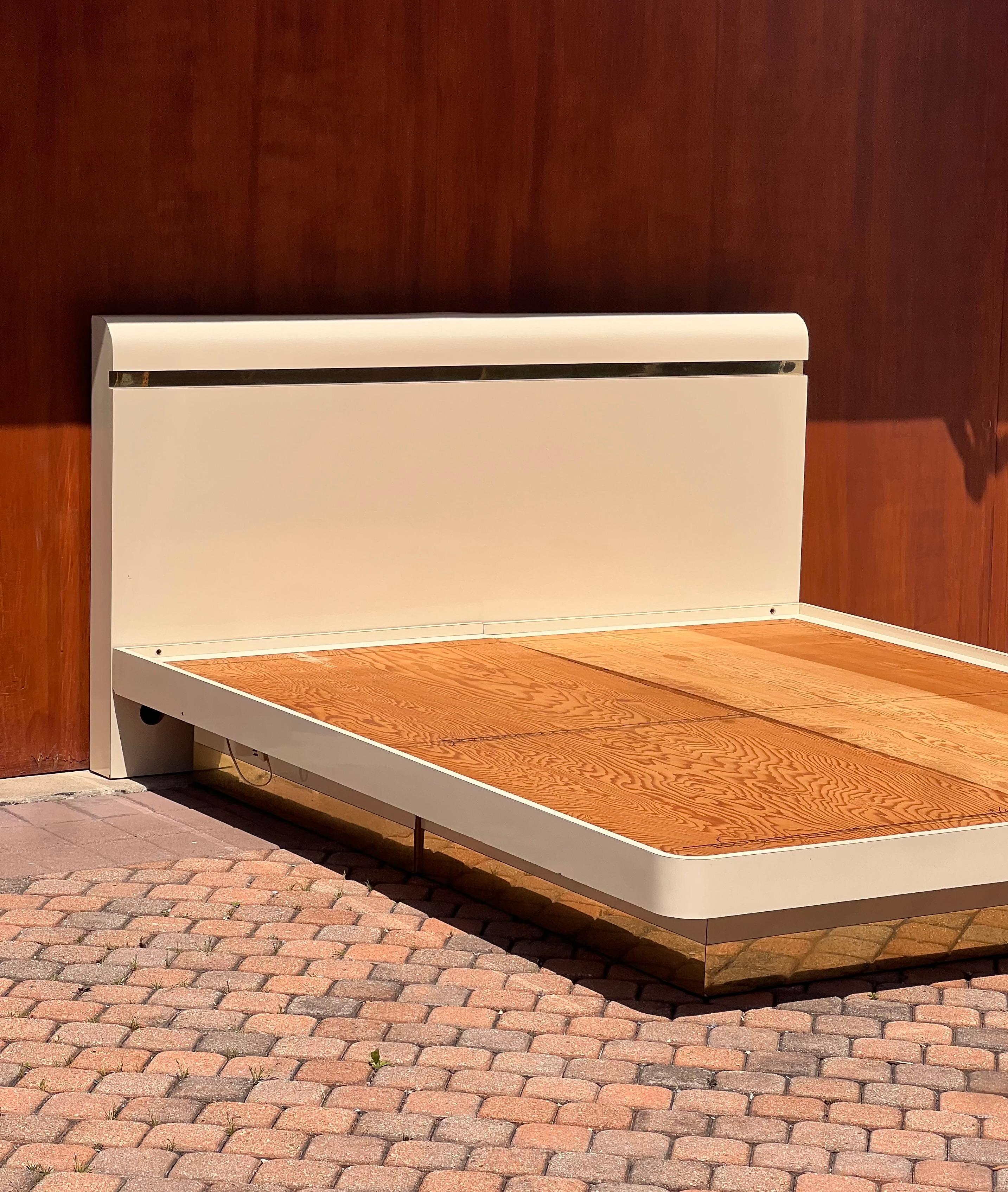 An Art Deco revival / Hollywood regency cream lacquered king bed frame with golden detail by APA, 1980s. This stunning piece, complete with curved geometry, gilded base and detail, and light-up underframe, will indubitably add glamour and flair to