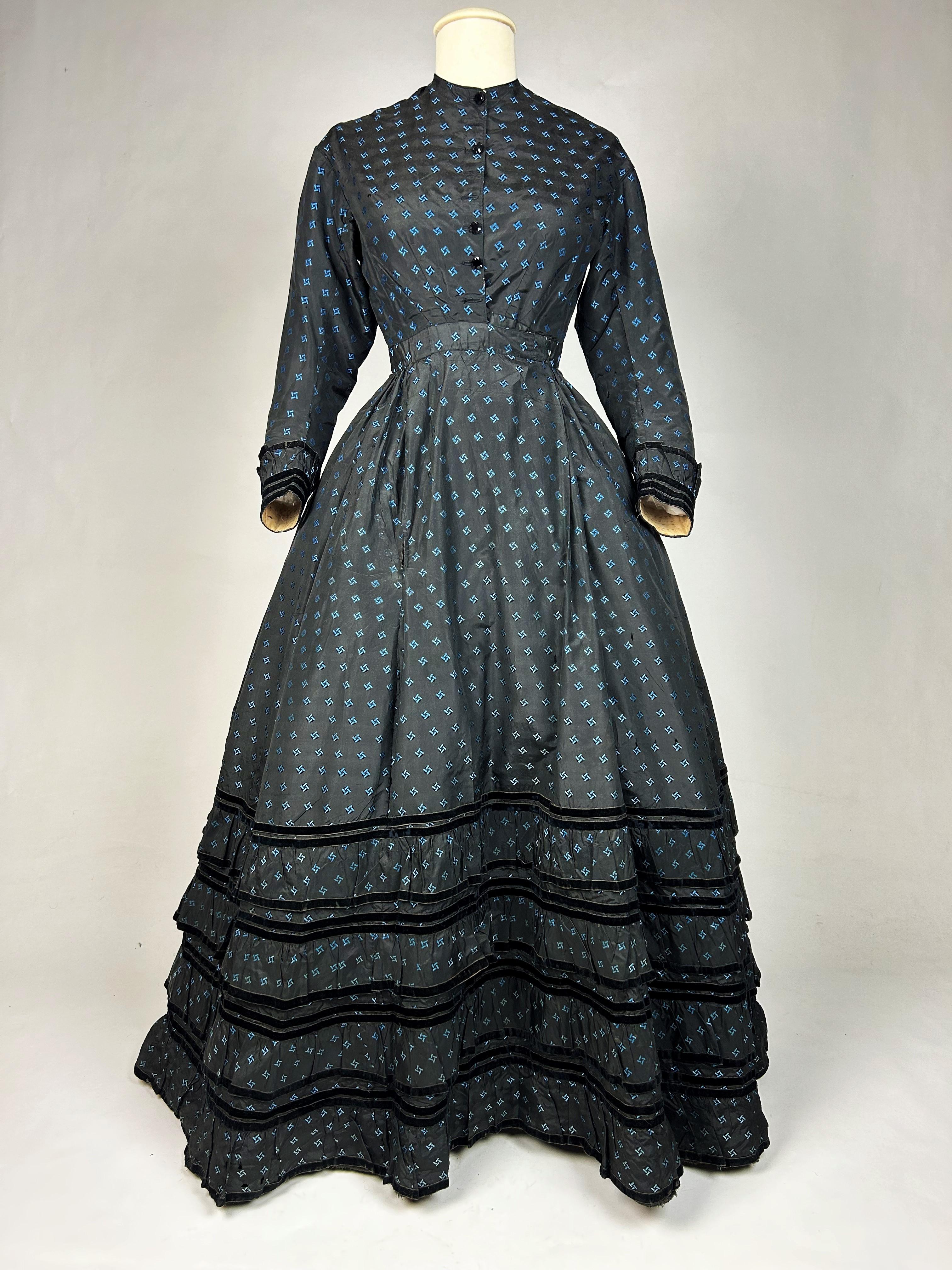 Circa 1870
France

Elegant Second Empire Crinoline day dress in black brocaded taffeta edged with blue geometric diamonds. Fitted bodice, crew neck, bent sleeves and front closure with black glass buttons (four buttons missing). Skirt with large
