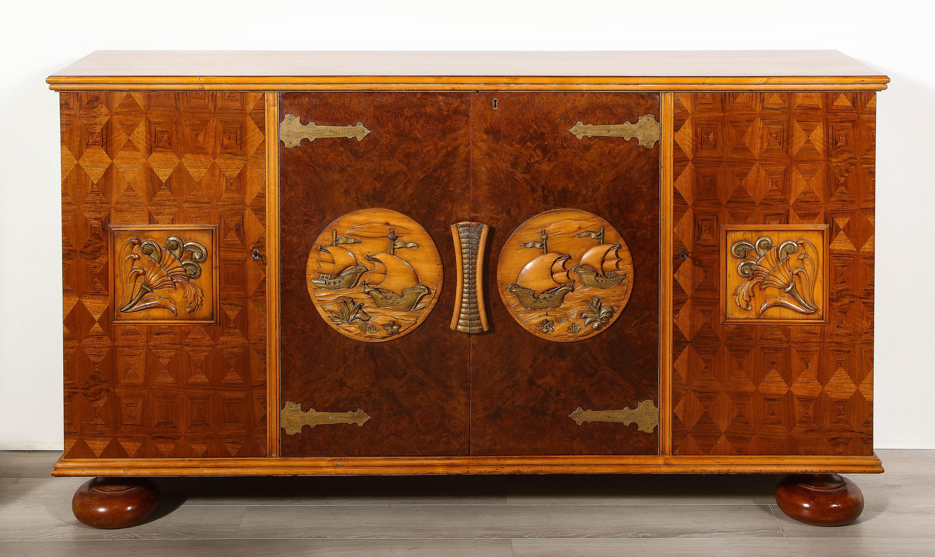 The custom sideboard dating from the 1950's, having a walnut veneered top over 4 doors each with a carved nautical scene relief. The outer doors and sides having parquetry veneer, the interior doors with burl walnut veneer. The interior itself fully