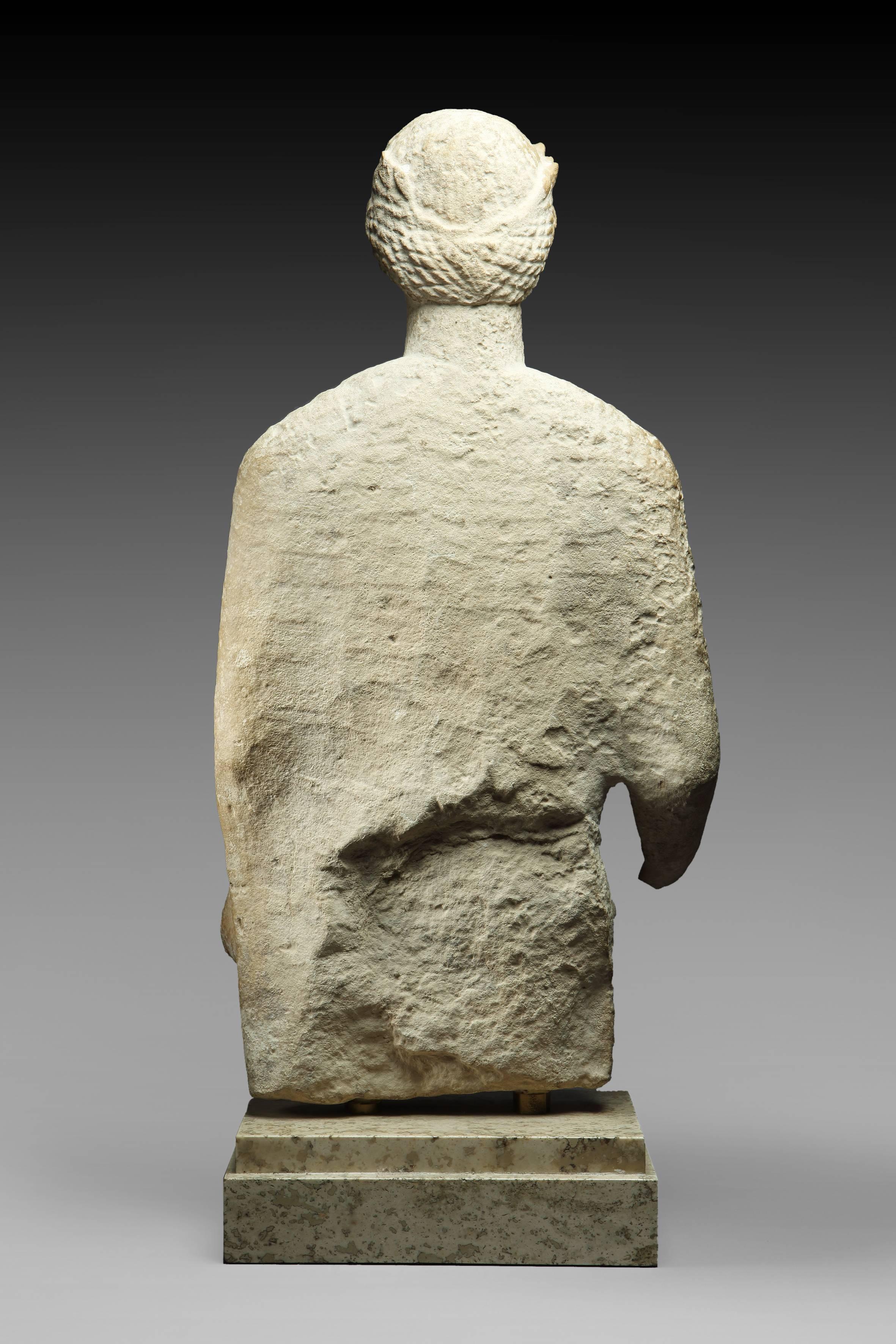 Represented frontally, his head is surrounded by a crown of laurels. He wears a tunic with fine folds held by a belt, a himation and holds in his left hand an object difficult to identify. Facial features, especially the smile, denote a deep
