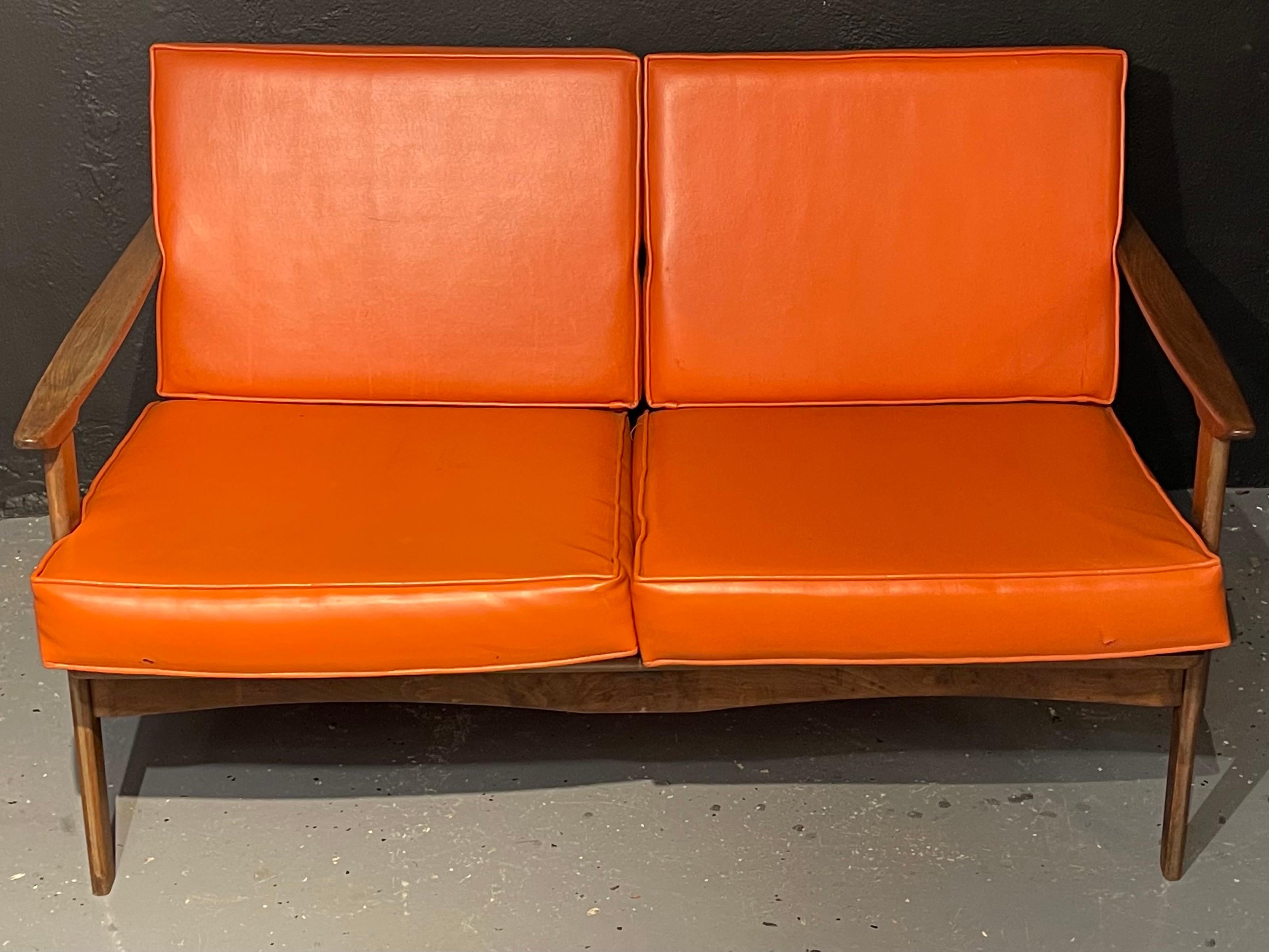 A Danish Mid-Century Modern settee having orange vinyl cushions in the manner of Ib Kofod-Larsen. All original. Slopping arms and sculpted skirt. The backrest of ladder form wooden slats with straps cushion seat rest supports. The whole of teak wood