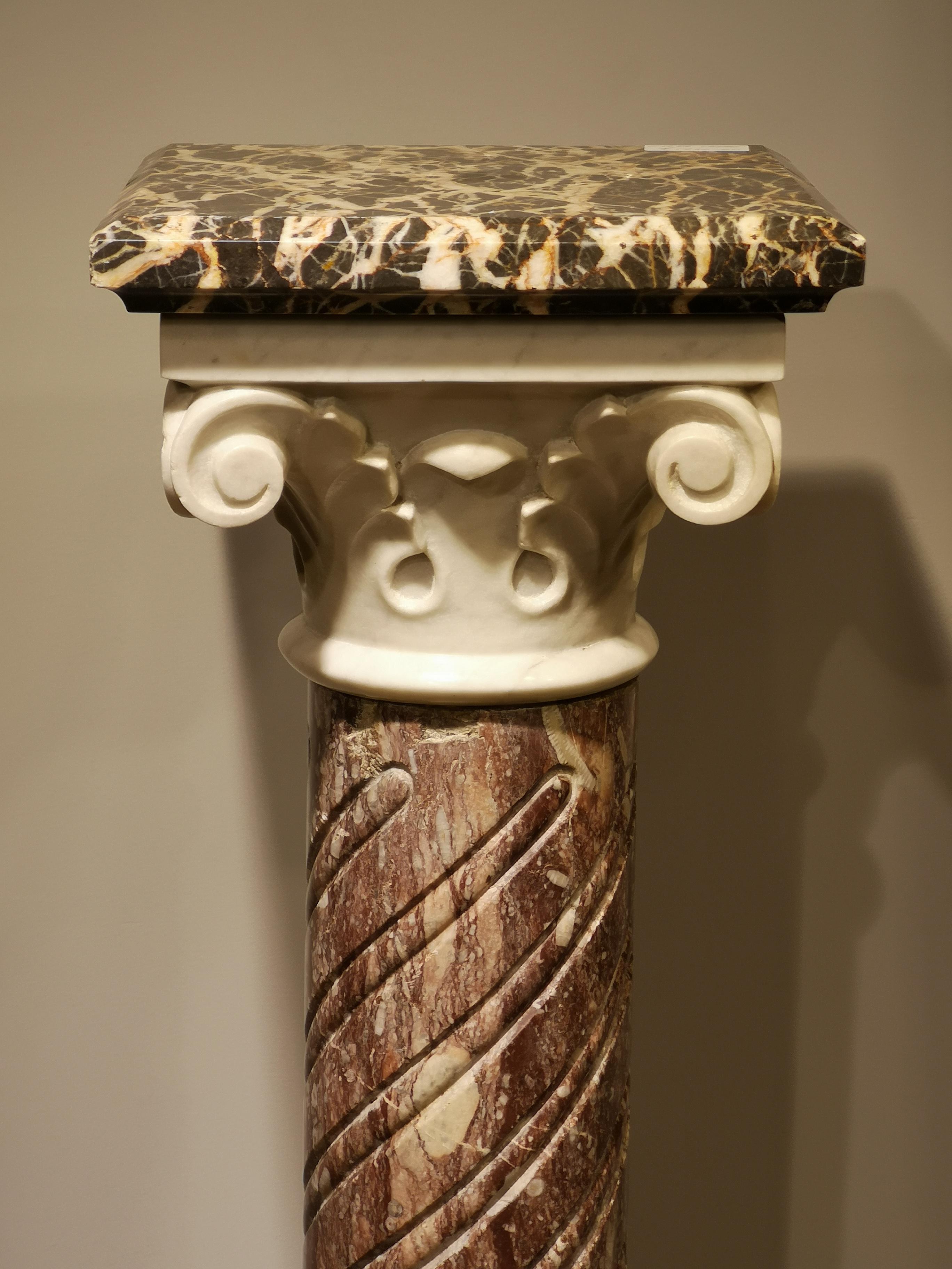 A decorative multi faceted column in the Gothic manner, the top of highly figured marble above white Ionic style volutes or scrolls and supported by a swirled and fluted narrow column, the whole on a decorative figured marble base with a collar of