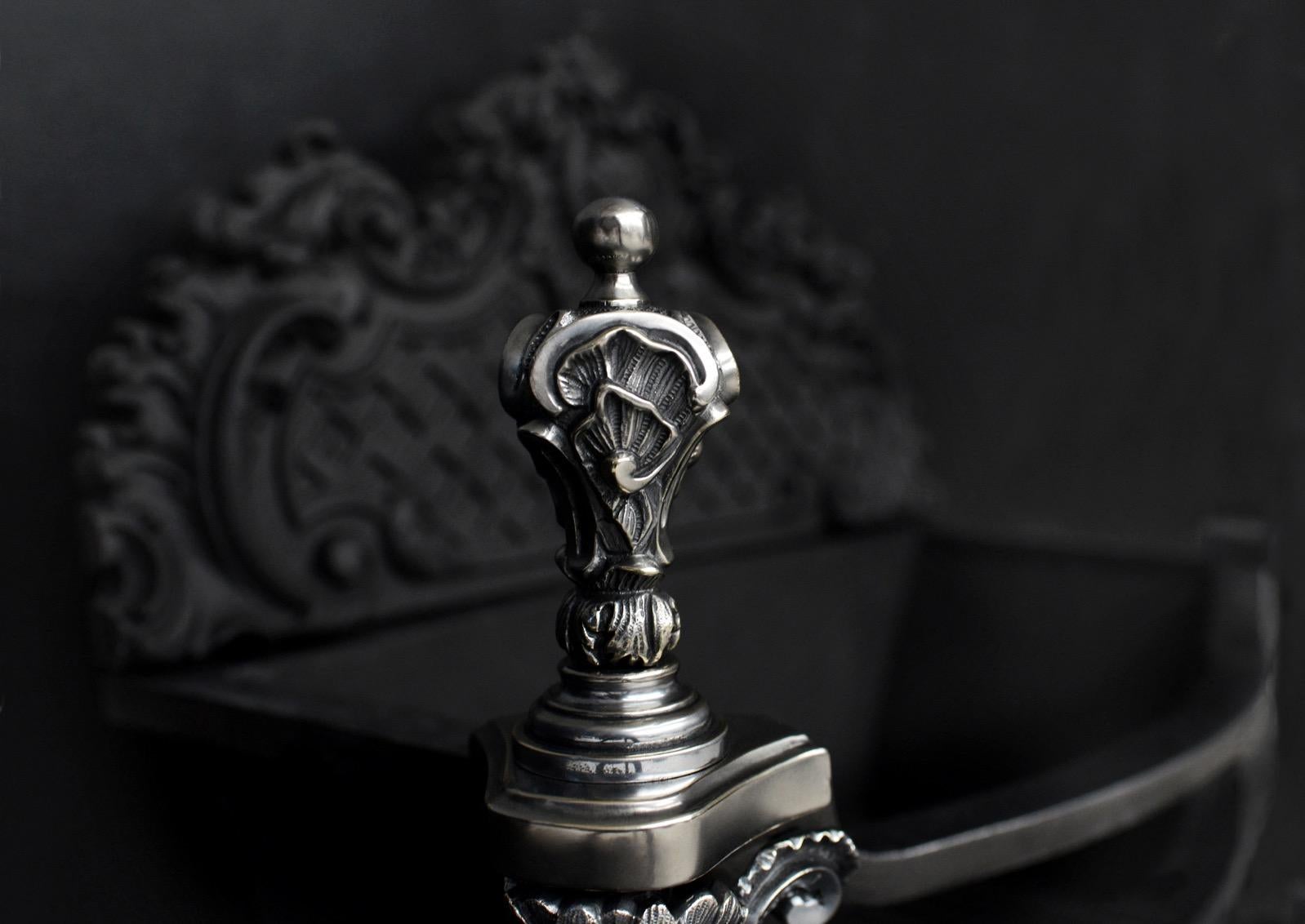 An elaborate Rococo firegrate in German silver. The ornately cast fret featuring foliage and shells. The legs with masks to sides, surmounted by decorate finals. The cast iron fireback behind with lattice and scroll work throughout. Circa 1900.