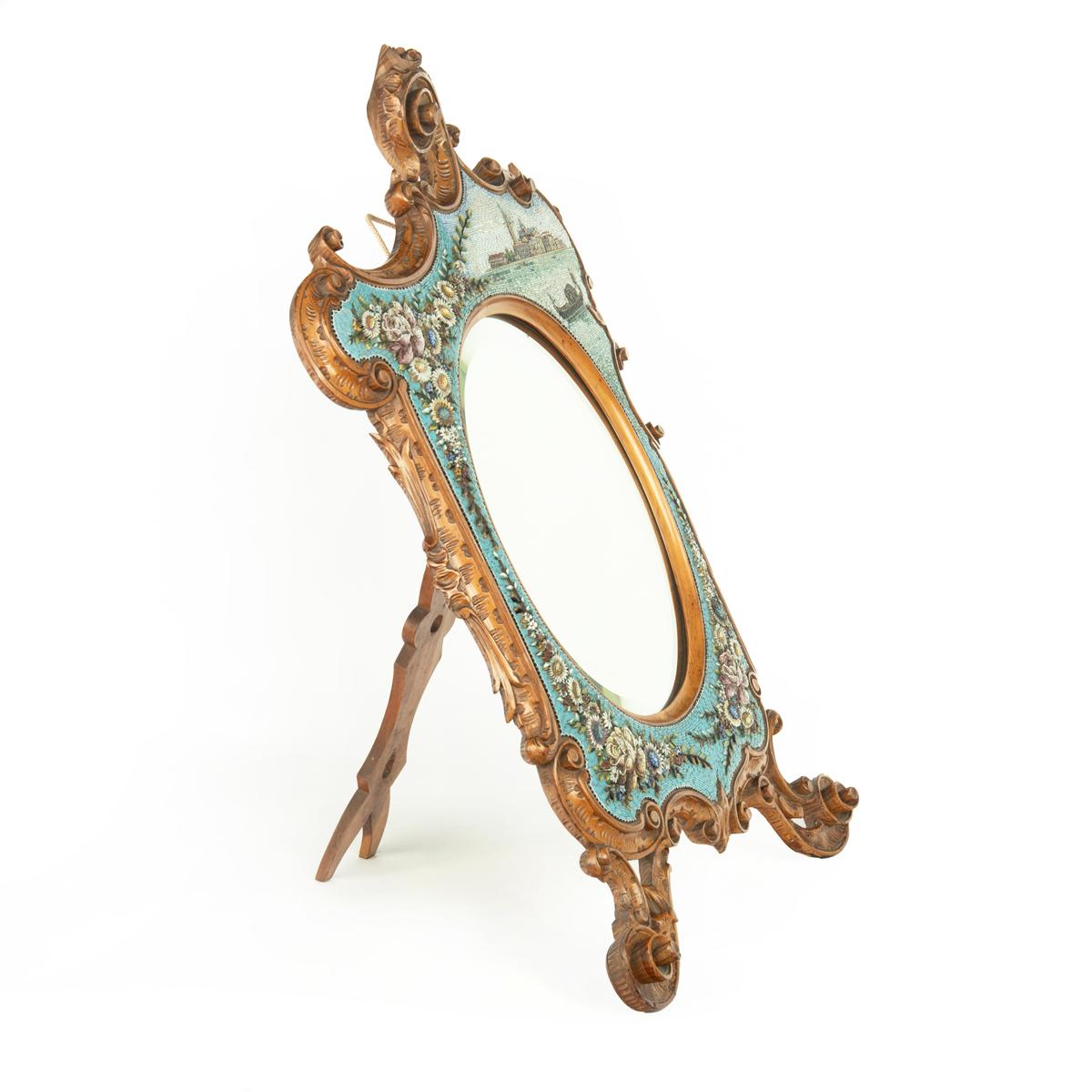 A delicate walnut easel dressing table mirror, the oval plate within an asymetrical frame of C-scrolls in the Rococco manner, .deocorated in glass mosaic with a Venetian scene and floral sprays. Italian, circa 1900.