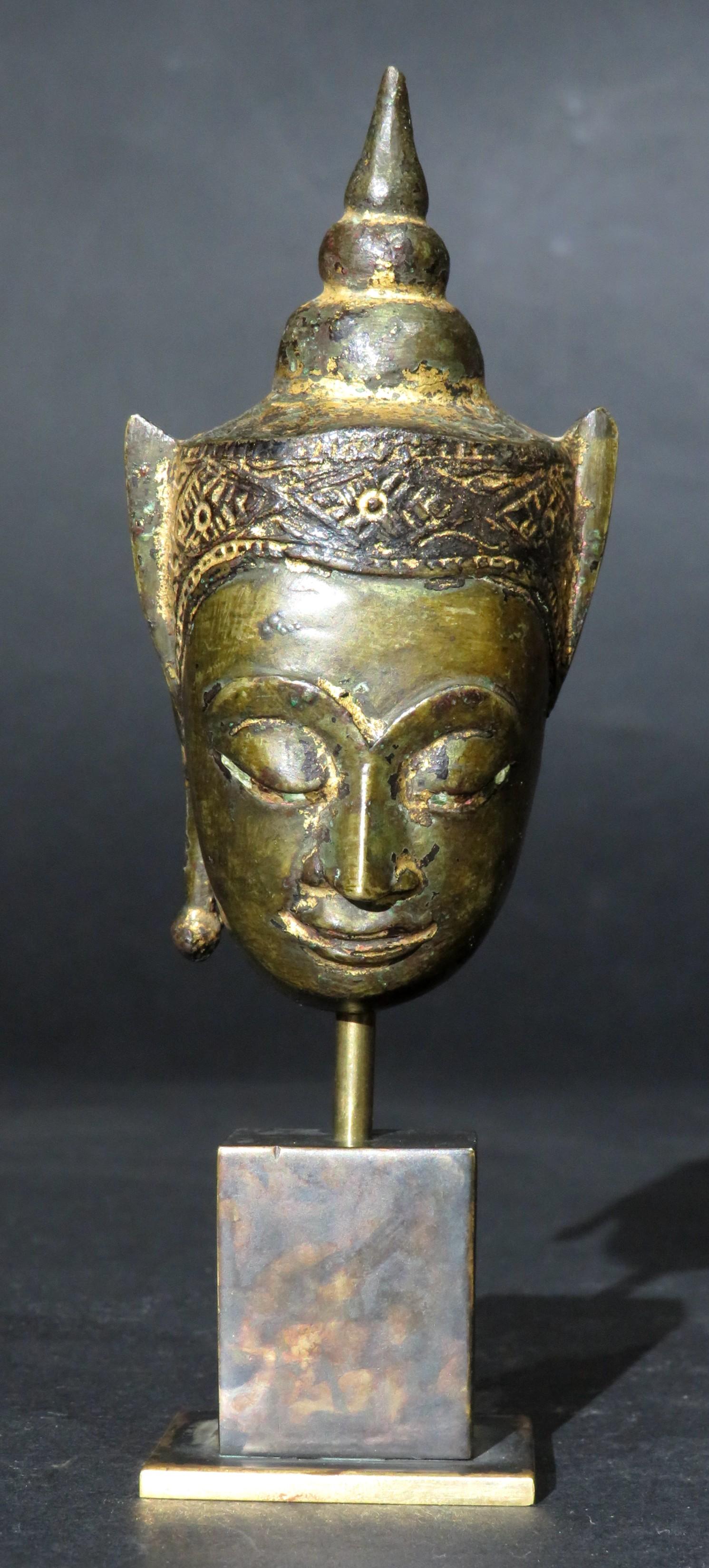 This 17th century Ayutthaya Period copper alloy (bronze) head of Buddha depicted in the ‘Phra Chao Song Khruang’ manner (Lord Wearing Ornaments), showing Buddha adorned with an ornate & partially gilded crown / diadem rising to a domed ushnisha. 
