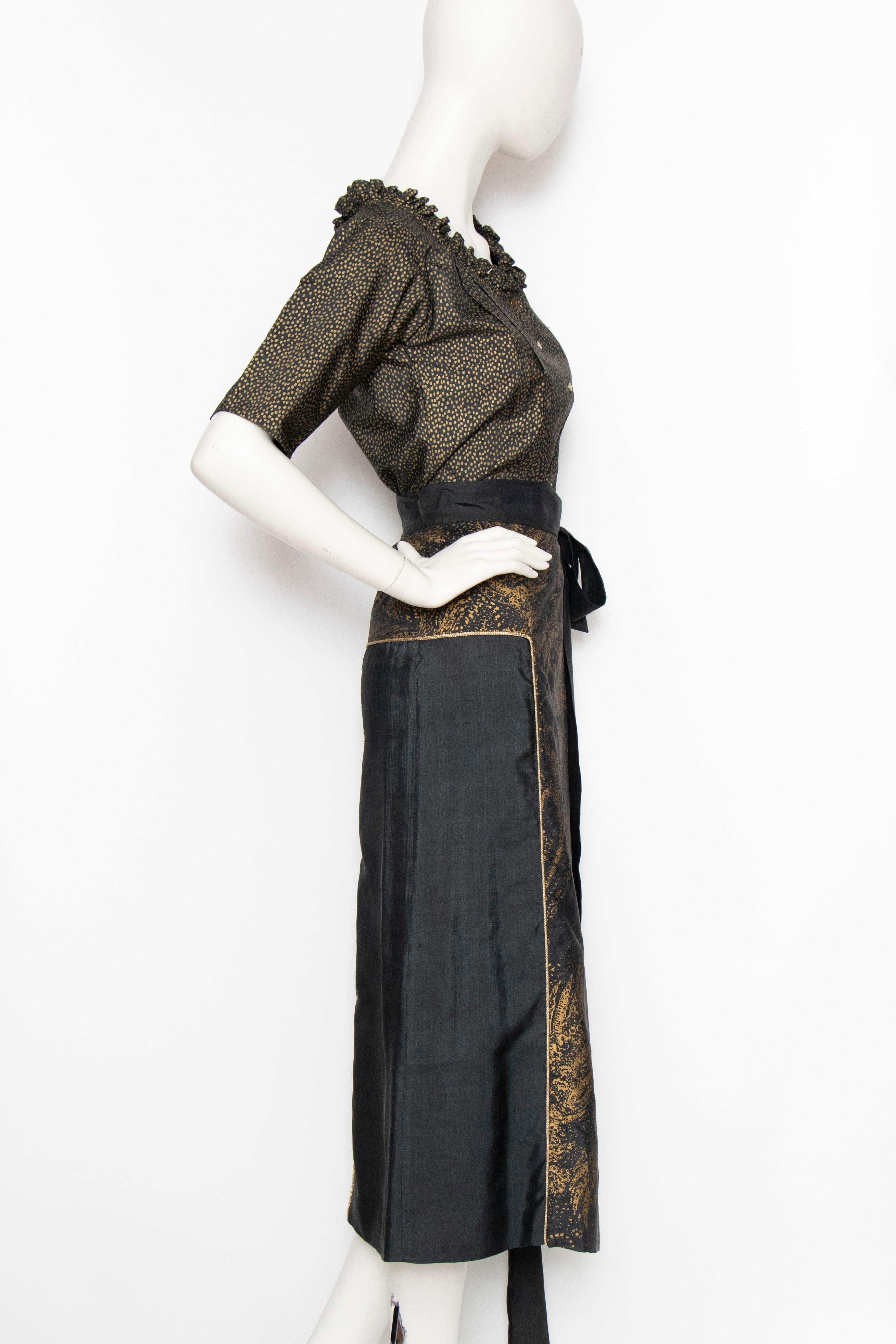 A 1980s Yves Saint Laurent Rive Gauche black and gold silk ensemble consisting of a short-sleeved blouse and wrap skirt. The blouse has a round neckline with small ruffle trim and a push-button front. The skirt has a gold paisley panel down the