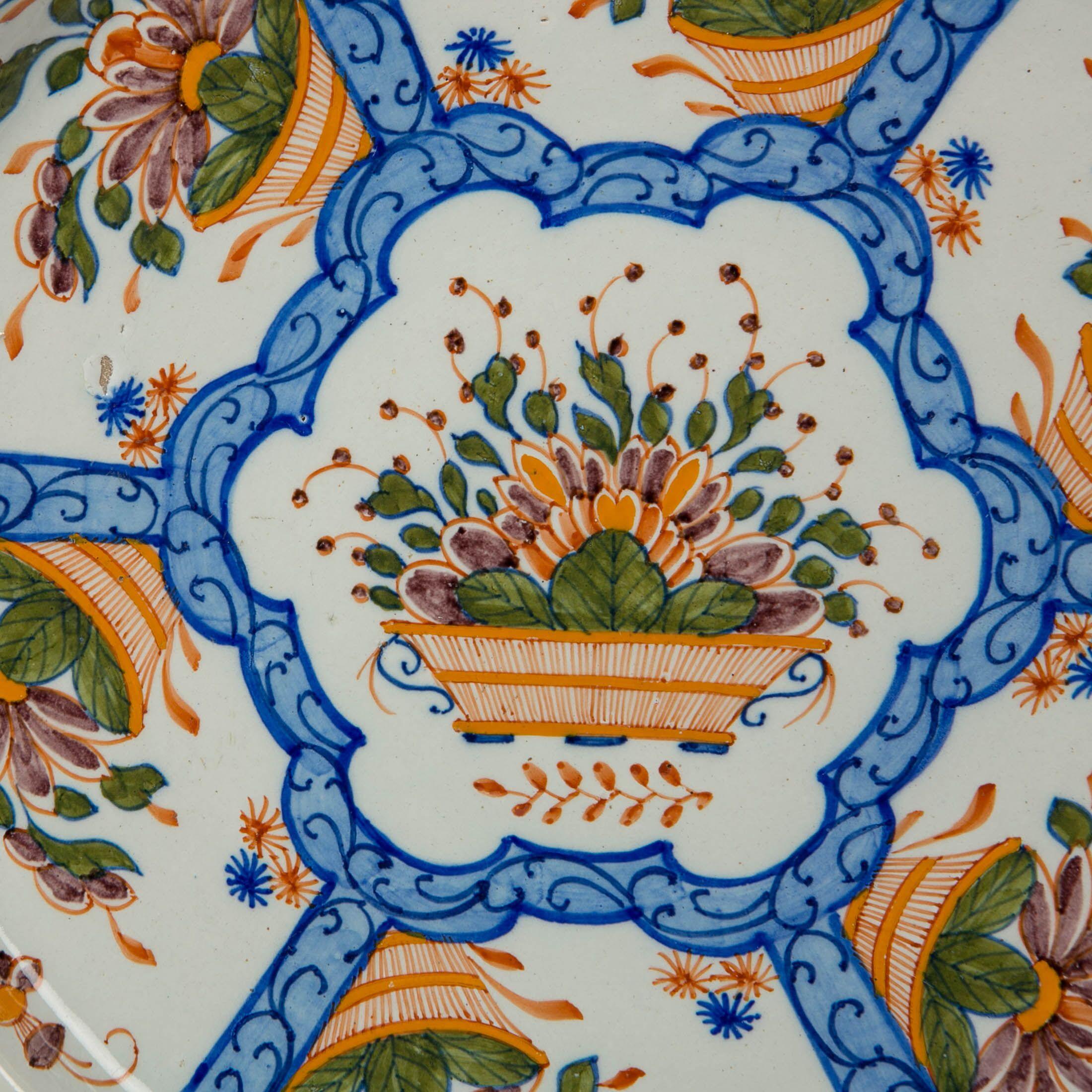 A Dutch Delft charger made in the early 19th century. It is painted with panels decorated with an orange flower basket overflowing with orange and manganese flowers, and green leaves. What jumps out visually are the light blue bands that divide the