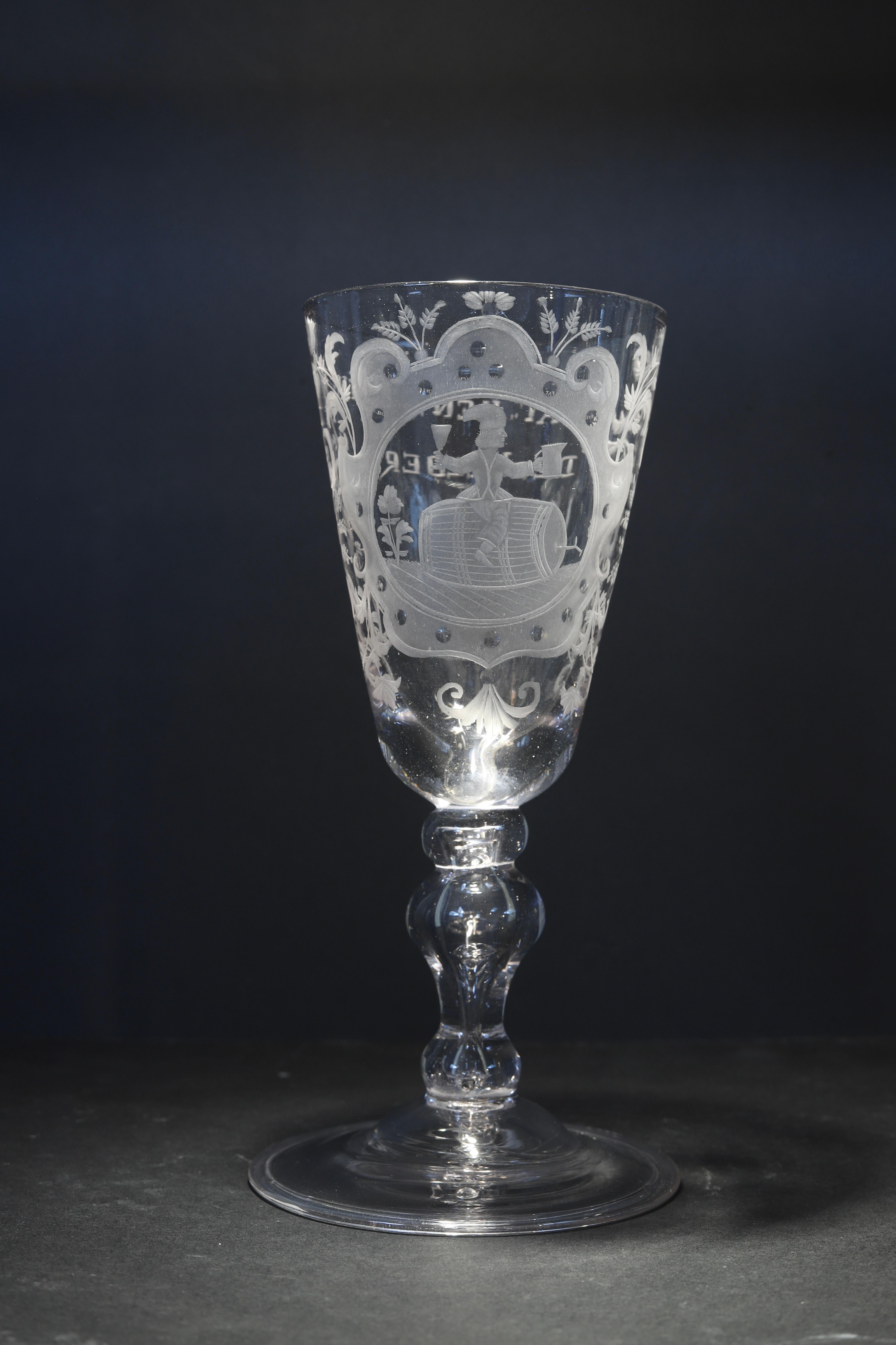 The Netherlands
Engraving: Dutch
Mid 18th century

A large Dutch engraved baluster wine glass, with a large funnel bowl with the decoration of a formal cartouche with a seated little guy on a wine barrel drinking and toasting, set on a thick stem