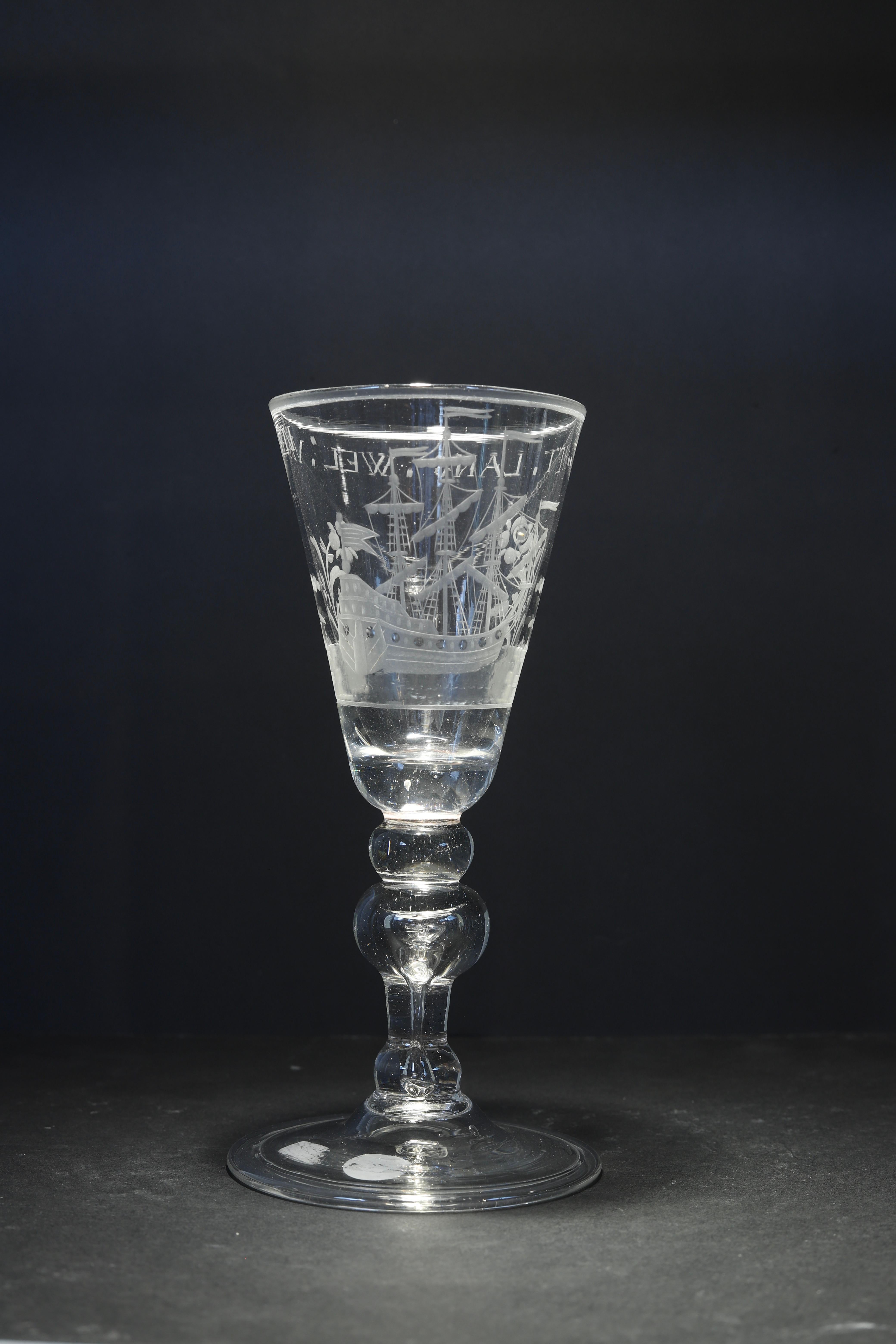 The Netherlands
Engraving: Dutch
Mid 18th century

A Dutch engraved baluster wine glass, with a tall pointed round funnel bowl with a solid base, decorated with a three-masted sailing ship at sea, flanked by stylized flowers, the stem with a