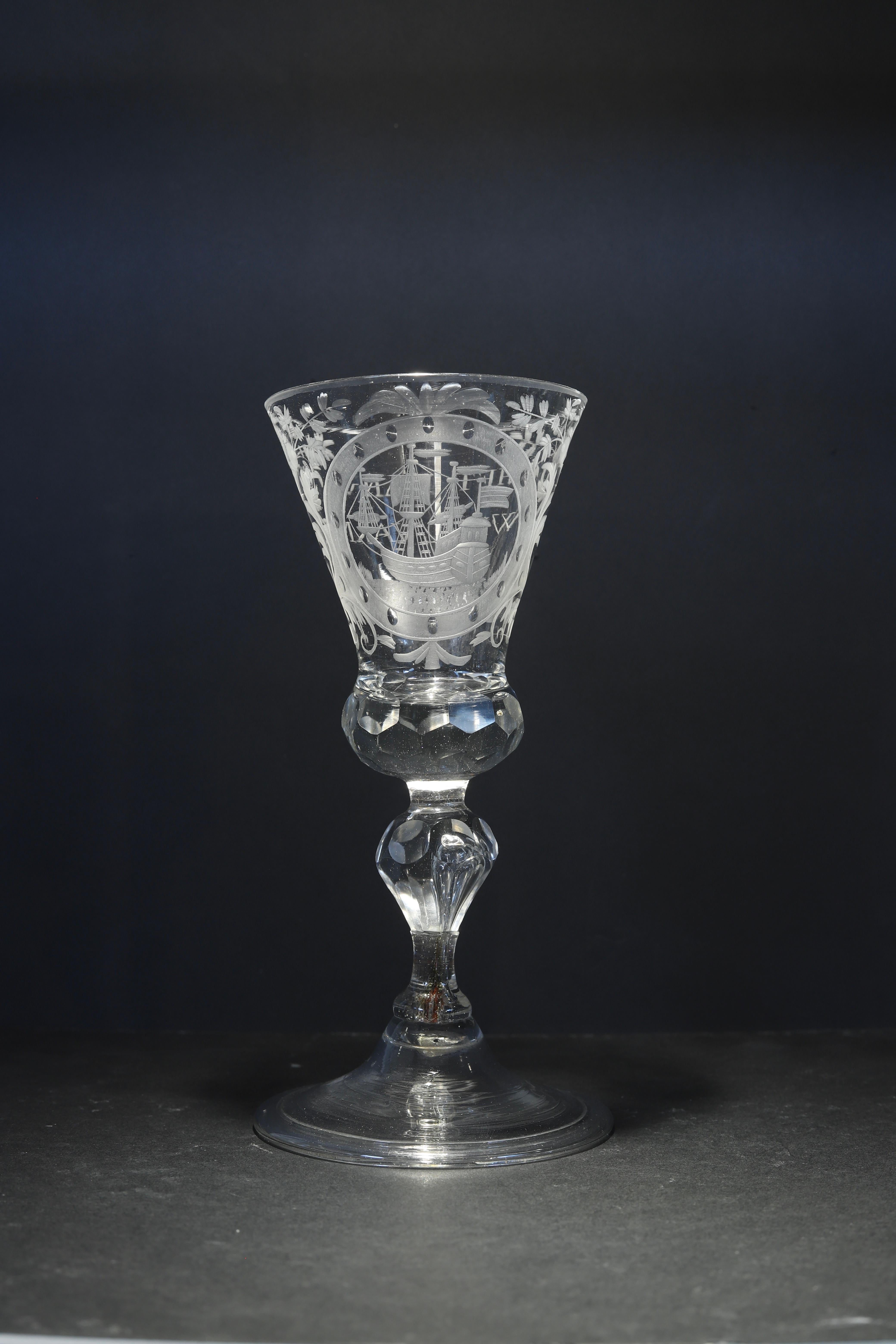 German, Saxon
Engraving: Dutch
Mid 18th century

A Saxon, Dutch engraved wine glass; the thistle bowl with a solid faceted base, decorated with a formal cartouche containing a three-masted sailing ship, flanked by scrollwork and floral sprays, on a