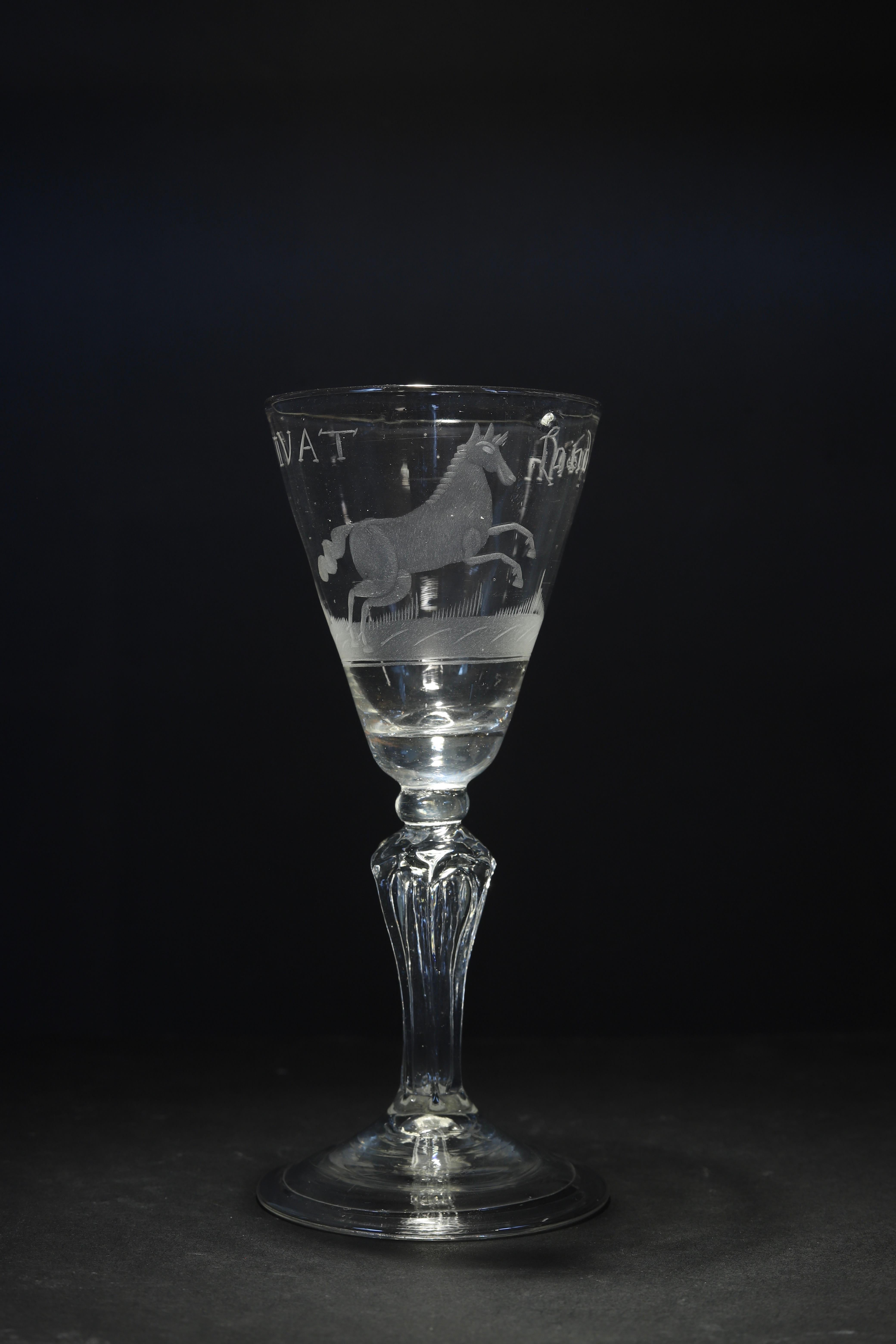 The Netherlands
Engraving: Dutch
Mid 18th century

A molded stem Dutch engrave wine glass with a pointed round funnel bowl decorated with the White Horse of Hanover rearing up on a sward, over a six-sided pedestal stem and folded conical