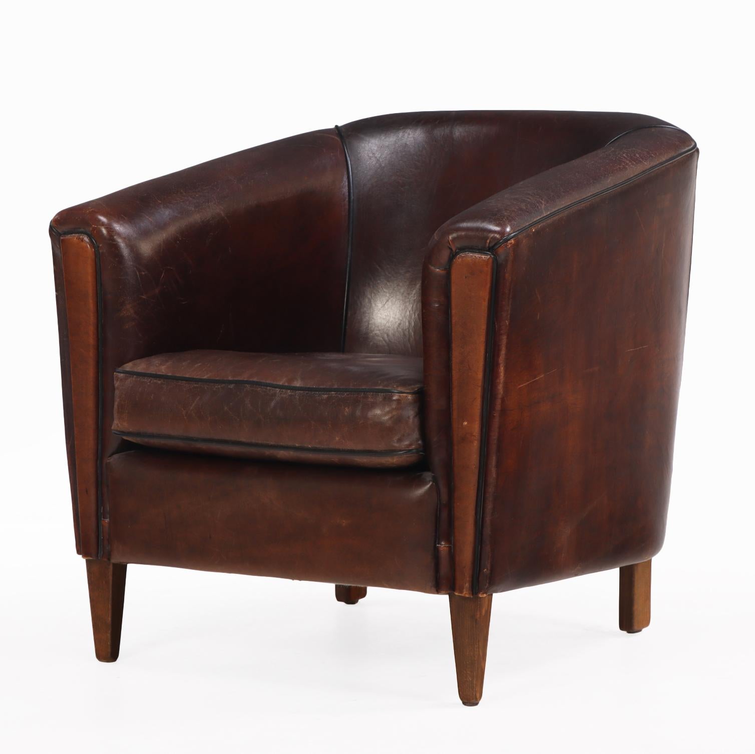 Ce fauteuil club en cuir sombre et luxueux présente une belle patine et des passepoils noirs contrastés. Il présente une silhouette classique aux proportions généreuses, avec une assise profonde et un dossier légèrement arrondi pour le confort et le
